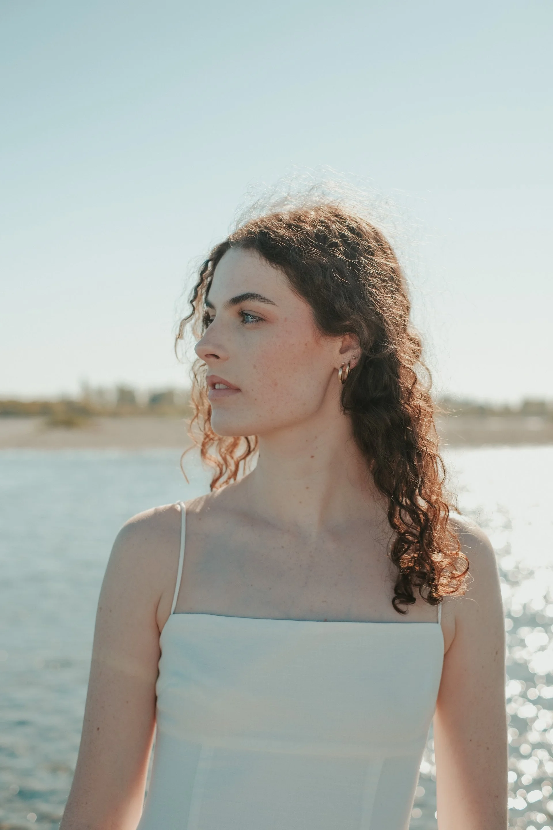 A woman with curly hair looking to the side outdoors near water on a sunny day.