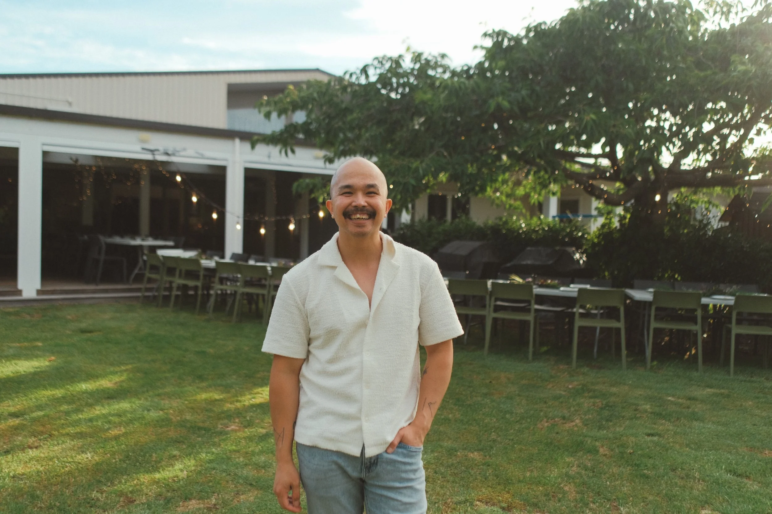 A smiling man with a mustache and a shaved head standing outdoors on a grassy area, with a building and trees in the background, during daytime.