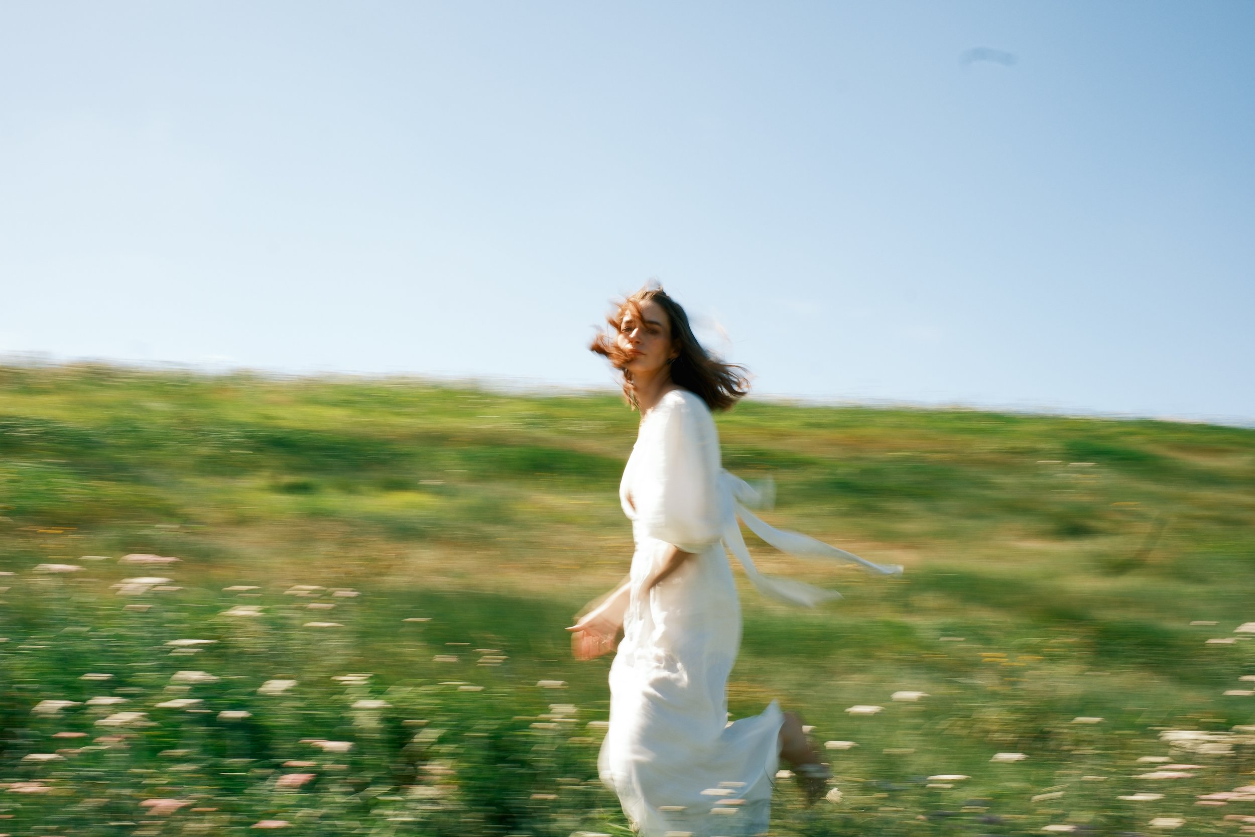 A woman wearing a white dress walking through a grassy field with a clear blue sky in the background.