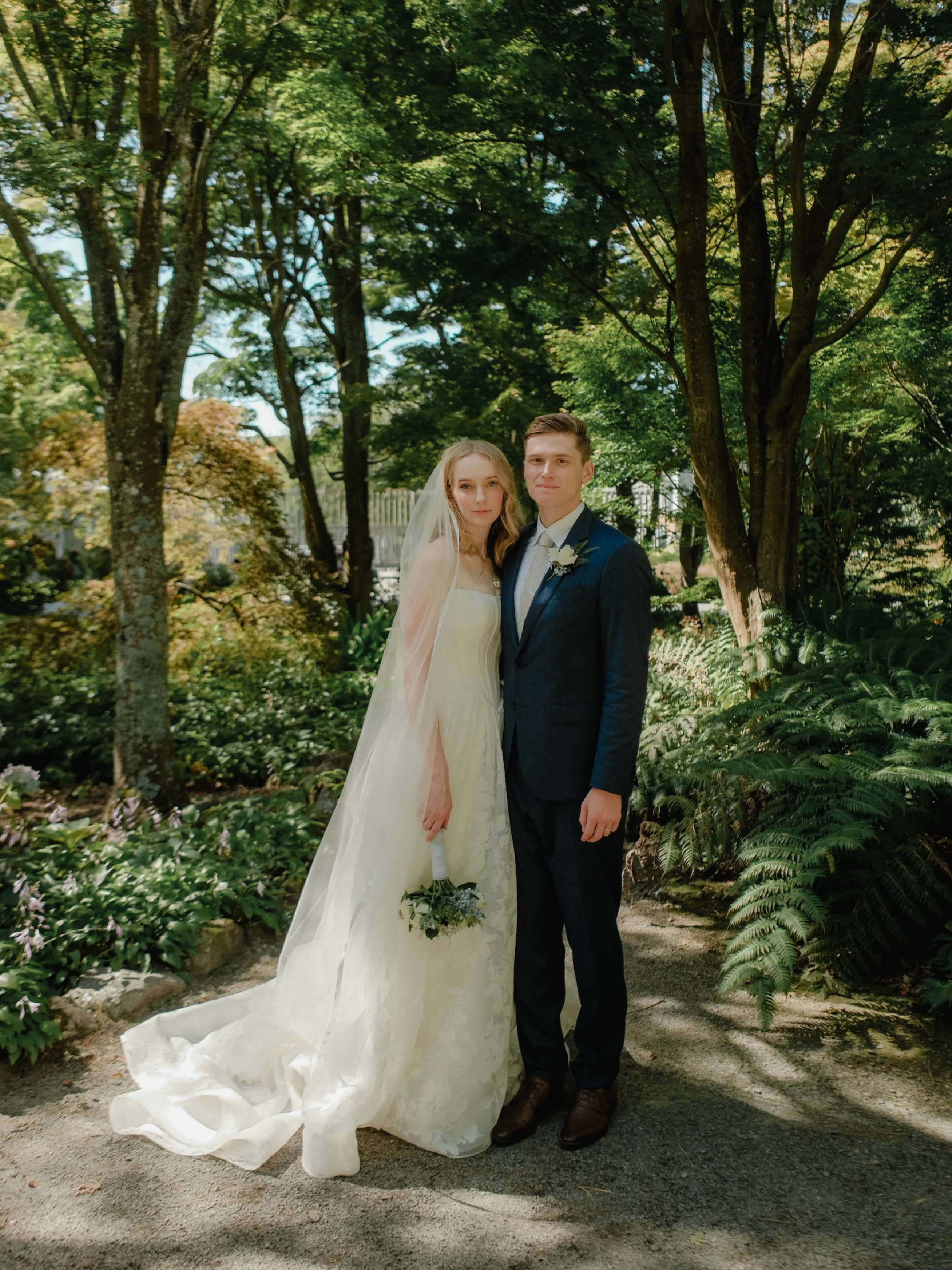 A bride and groom standing together outdoors in a lush, green forest setting for their wedding photo. The bride is wearing a white wedding gown and veil, holding a small bouquet. The groom is dressed in a dark suit and tie.