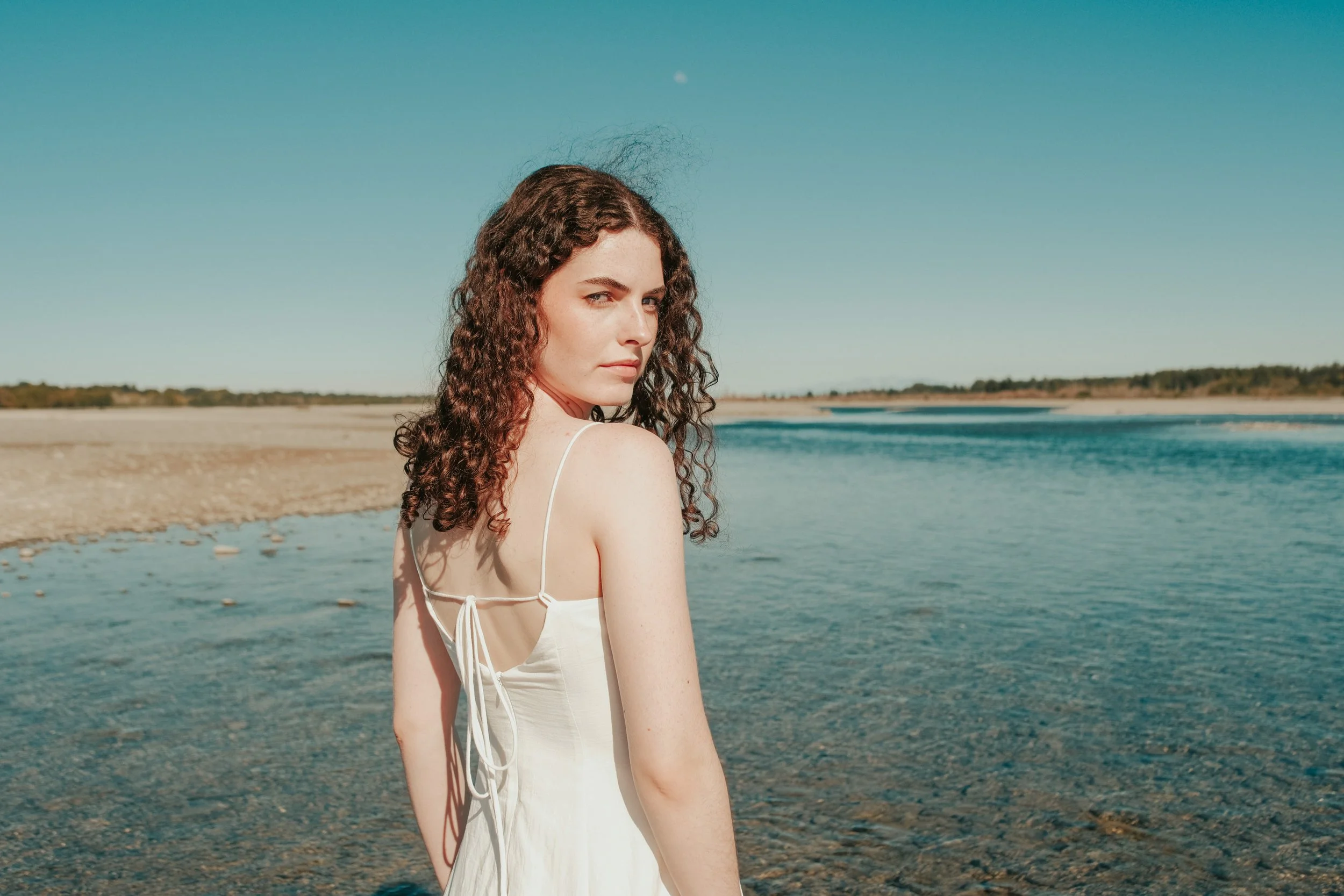 Young woman with curly brown hair wearing a white dress standing by a river during daytime.