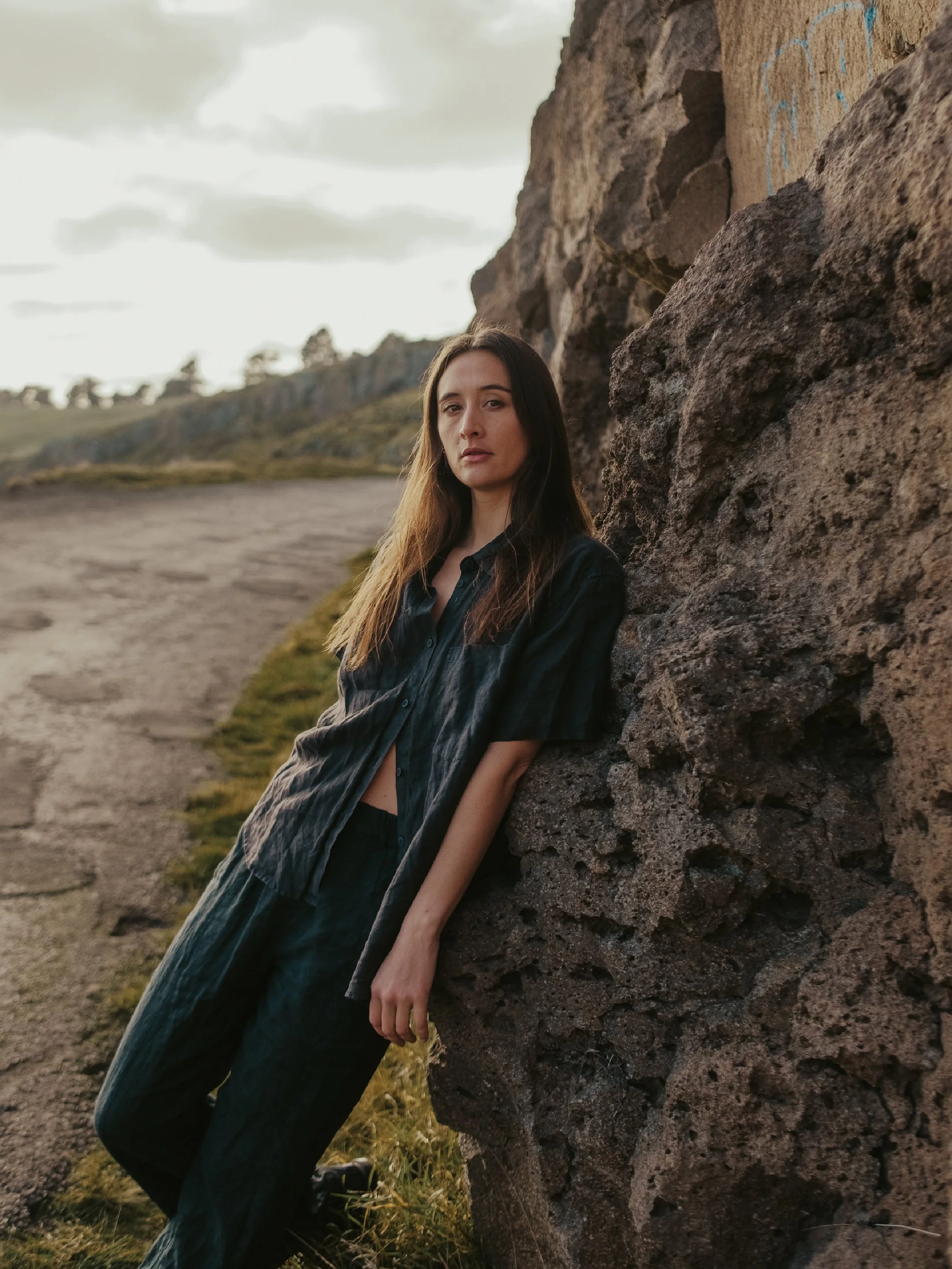 A woman leaning against a large rock outdoors with a scenic landscape of hills and cloudy sky in the background.
