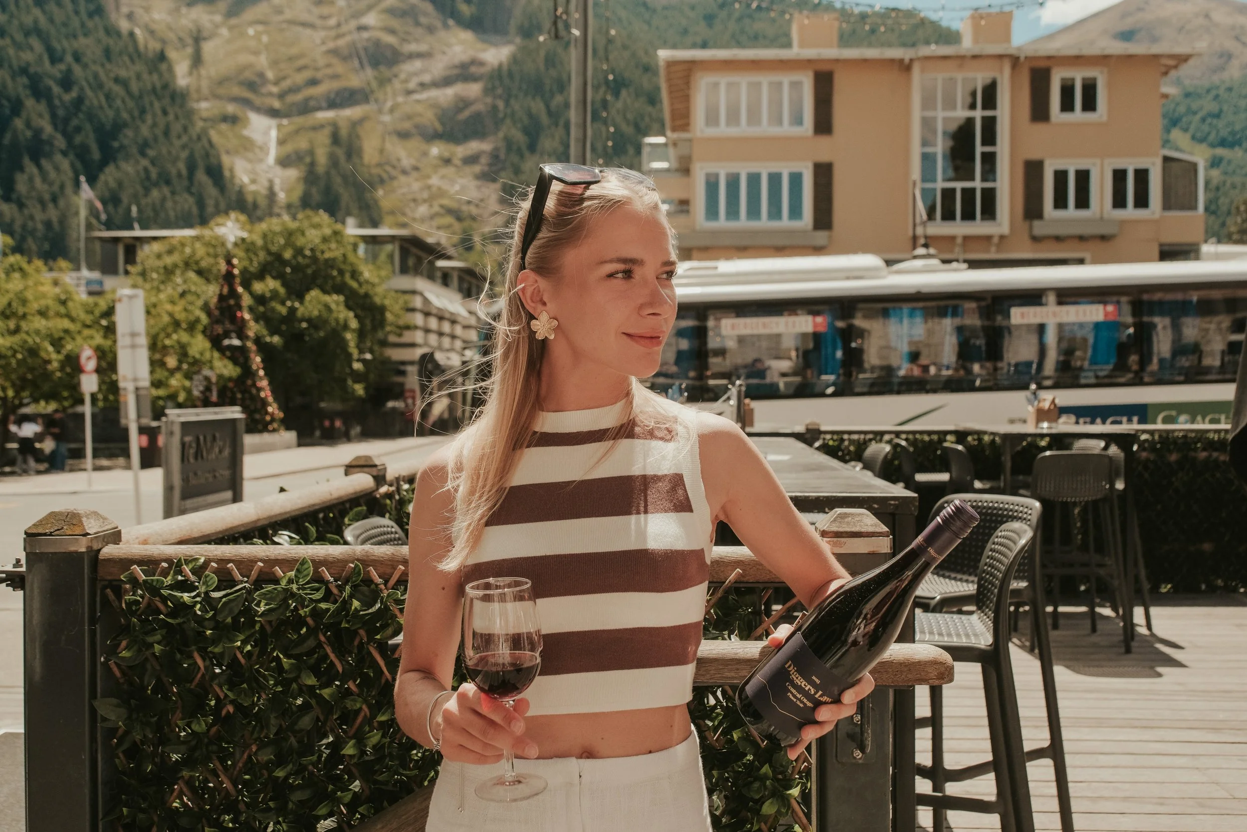 A woman with blonde hair holding a glass of red wine and a wine bottle, standing outdoors on a sunny day with mountains and buildings in the background.