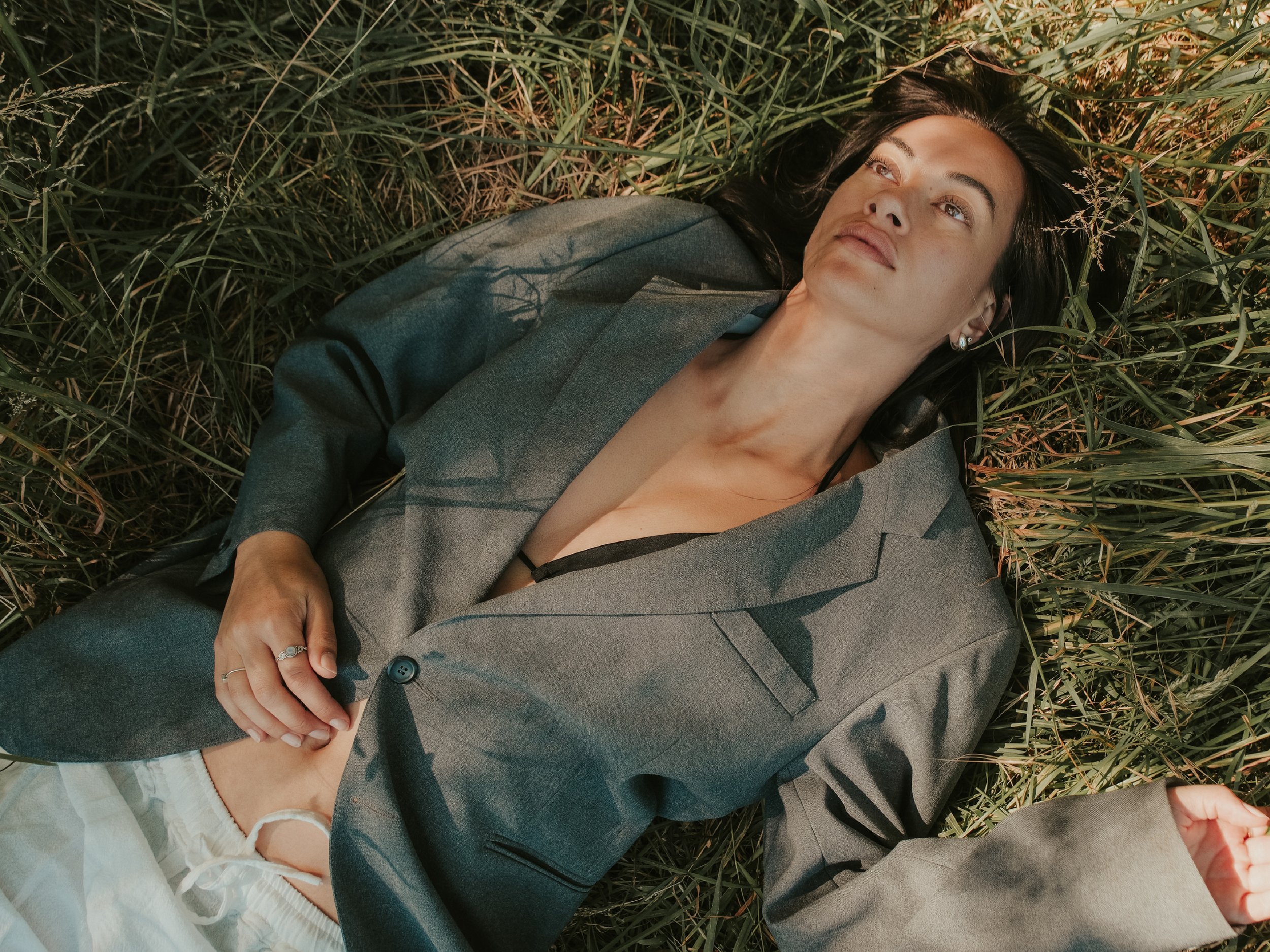 A woman lying on her back in a field of tall grass, wearing a gray blazer with a black bra visible underneath. She has dark hair, earrings, and a ring on her left hand, and is looking up with a thoughtful expression.