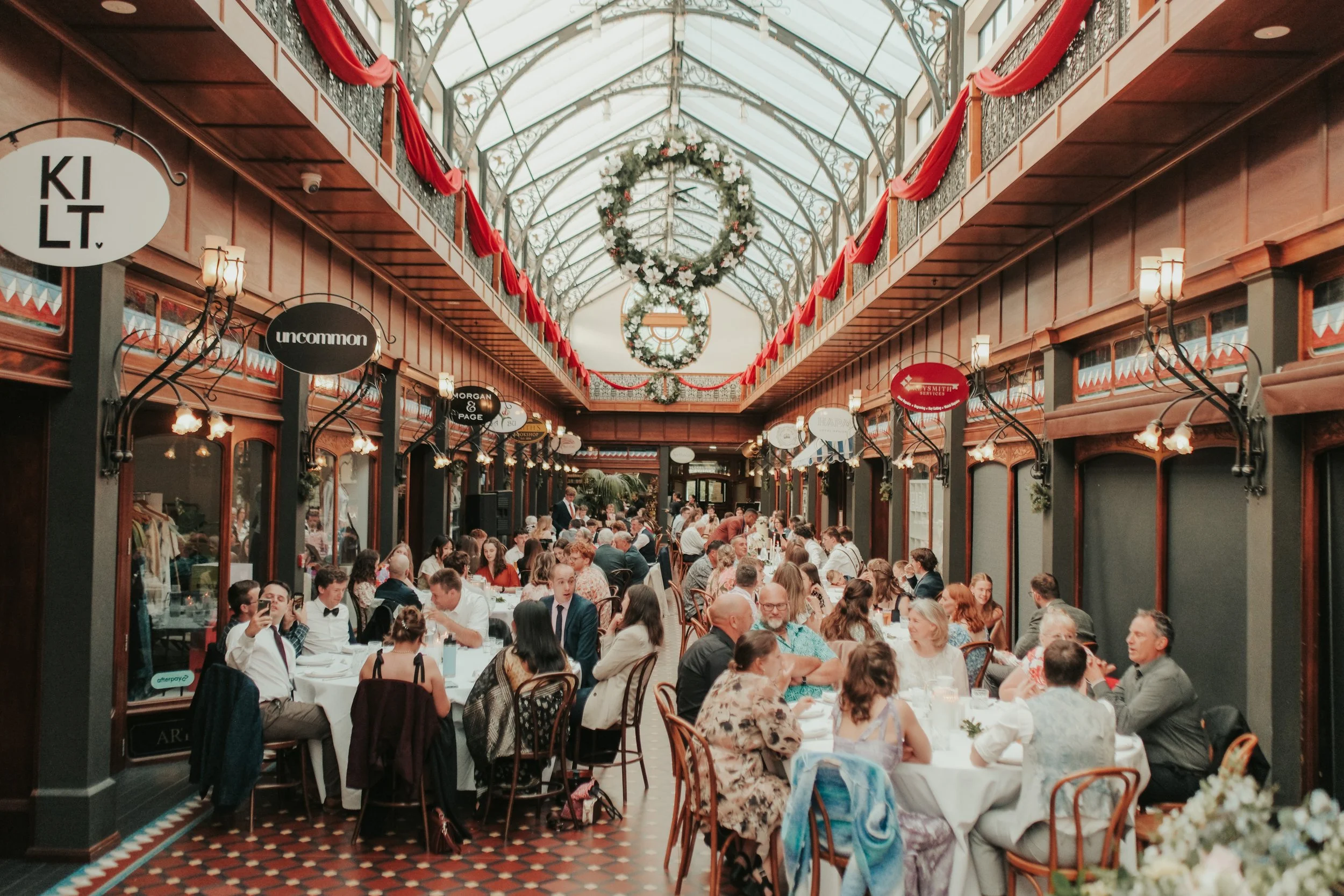 A busy indoor restaurant with a glass ceiling decorated for the holidays, featuring red drapes and wreaths. Many diners are seated at round tables enjoying their meals.
