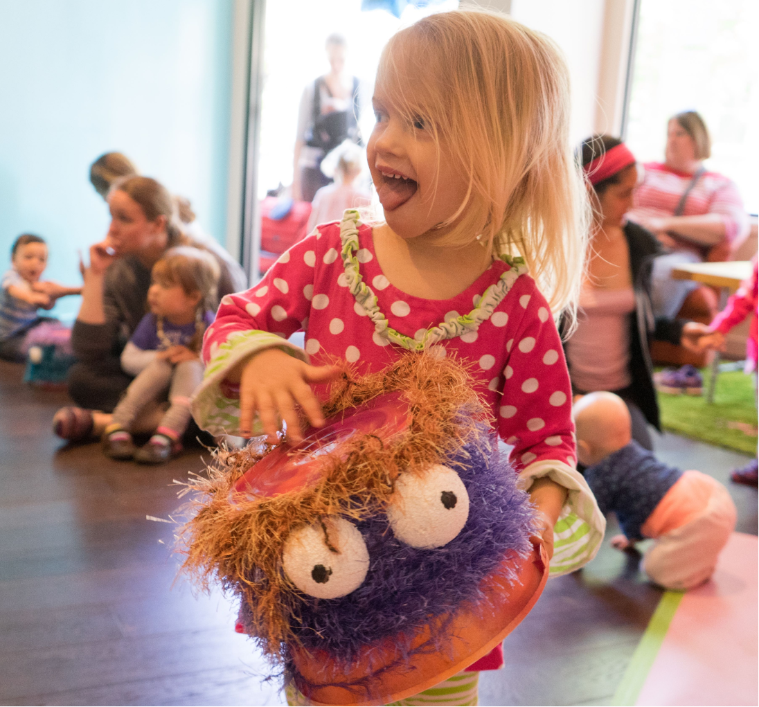 A young girl with blonde hair playing with a colorful puppet in a busy indoor play area, with other children and adults visible in the background.
