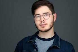 Portrait of a young man with glasses, dark hair, and a beard, wearing a denim jacket and a gray shirt, against a plain background.