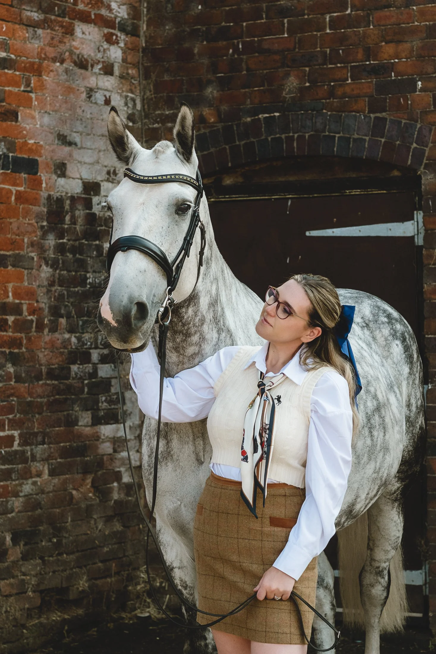 A woman wearing glasses, a white shirt, a cream-colored vest, and a brown plaid skirt stands next to a gray and white horse in front of a brick stable door.
