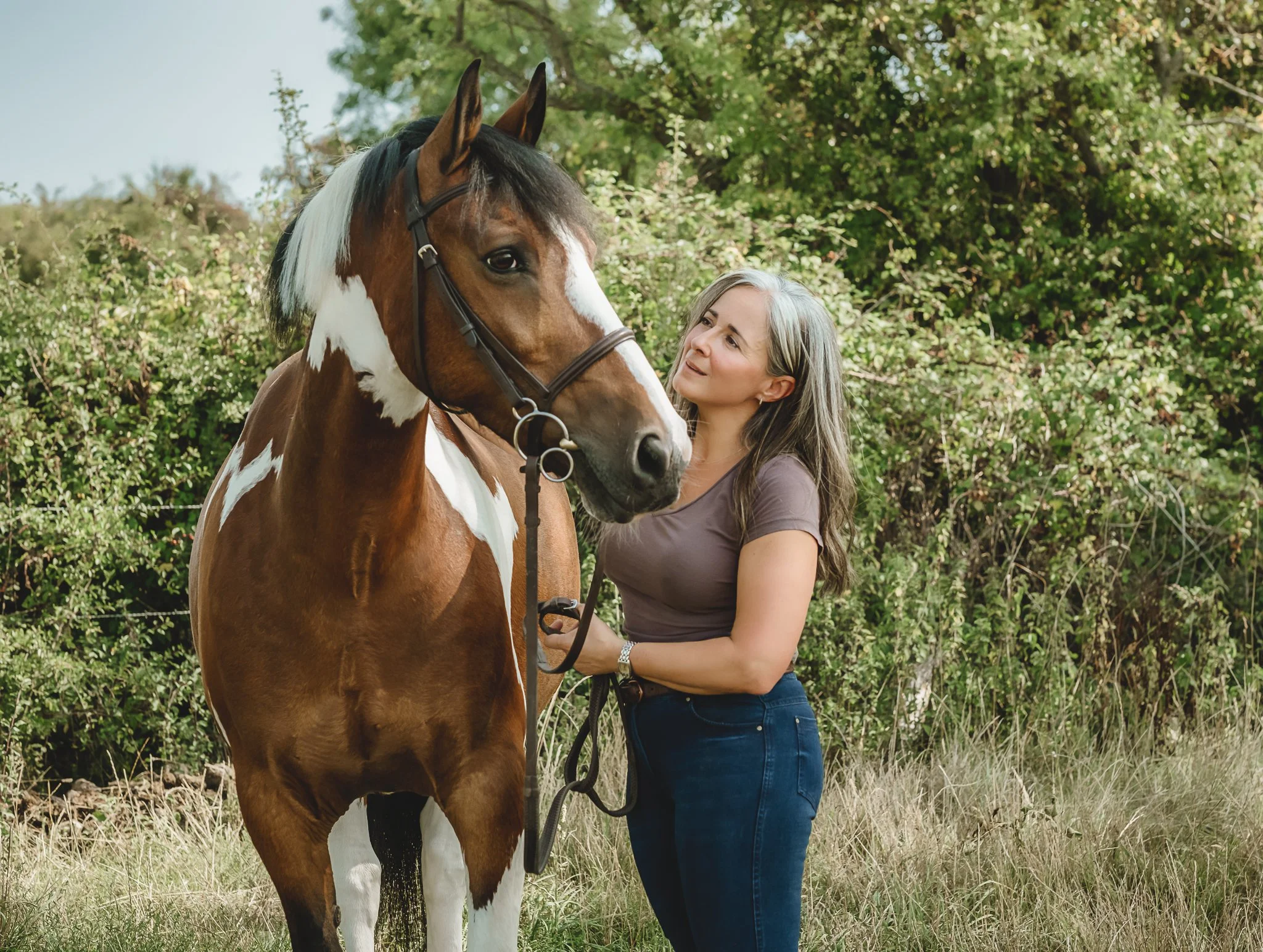Woman holding the reins of a brown and white horse outdoors with green trees in the background.