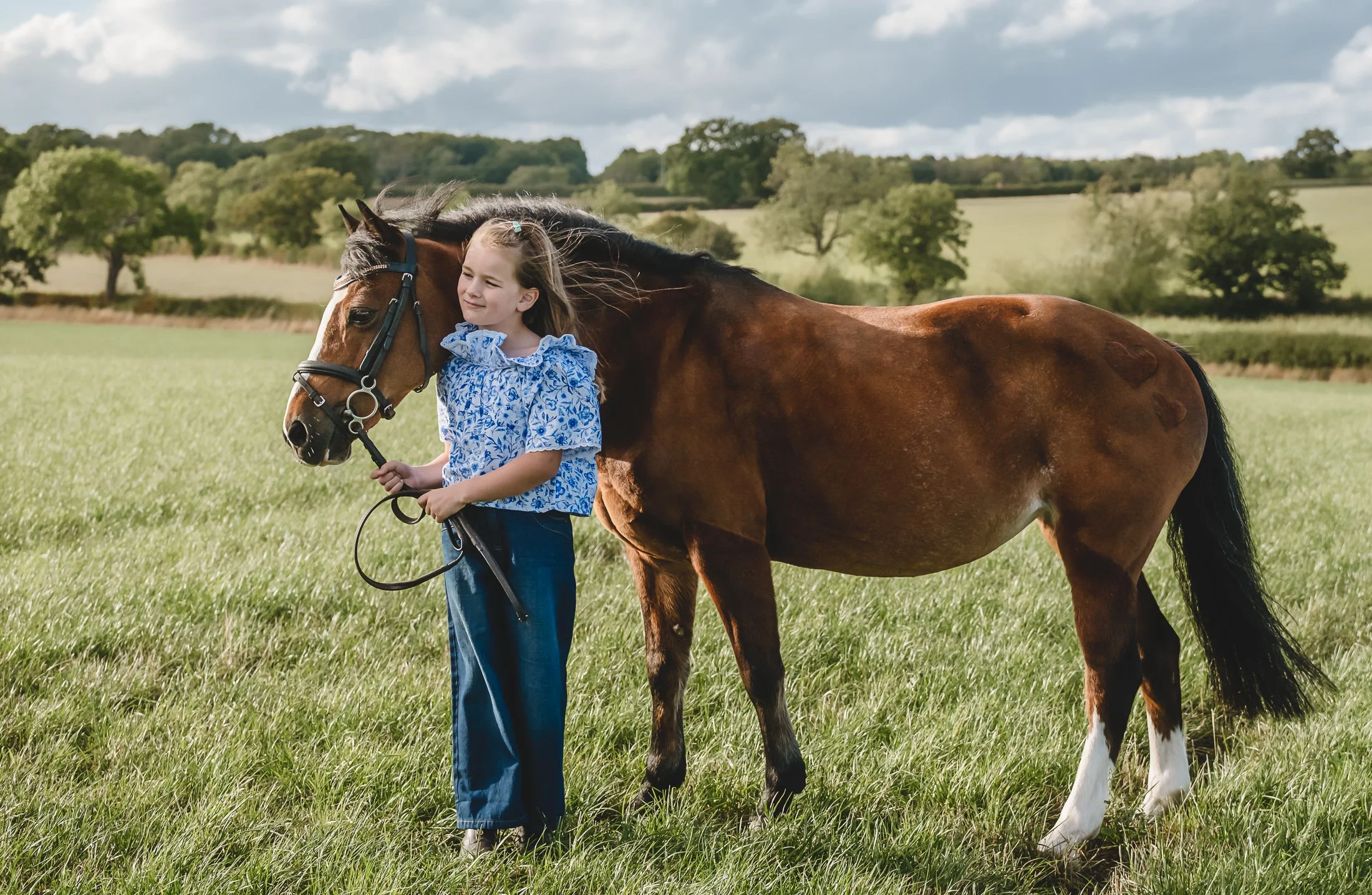 A young girl standing in a grassy field, holding the lead of a brown horse with black mane and tail. The girl is wearing a blue and white patterned blouse and denim jeans. The background shows trees, rolling hills, and a partly cloudy sky.