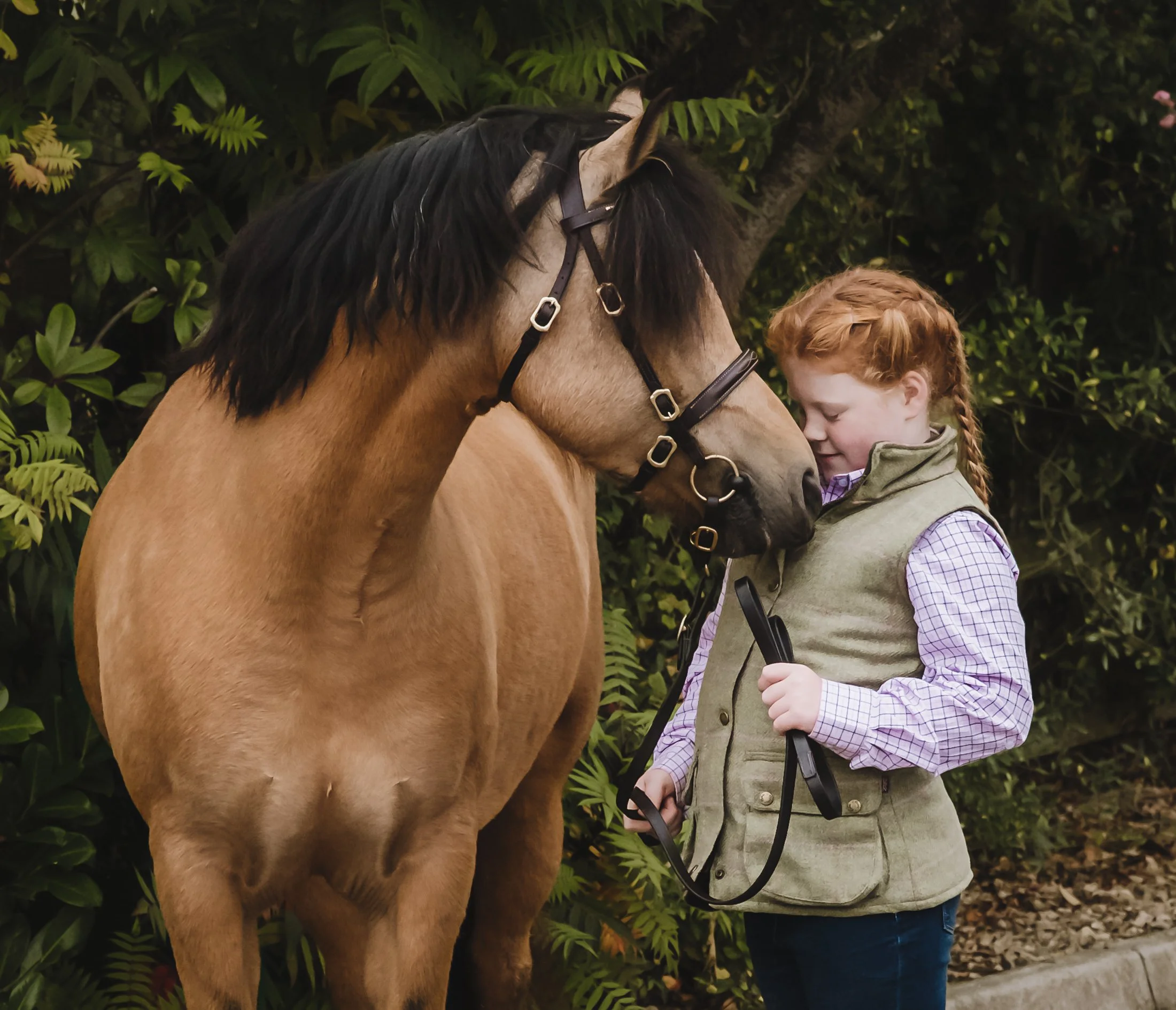 A young girl with red hair and a braid, wearing a vest over a plaid shirt, stands close to a light dun pony with a black mane, gently touching its forehead while holding the horse's lead rope, with green foliage in the background.