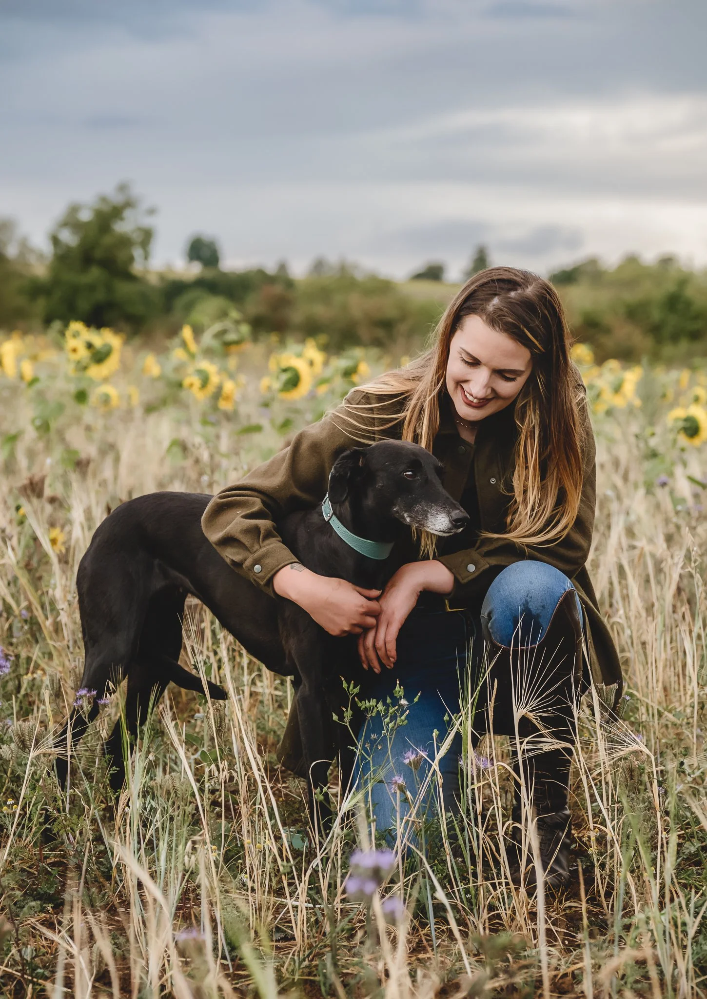 A young woman kneeling in a sunflower field with a black dog, both smiling and enjoying the outdoors on a cloudy day.