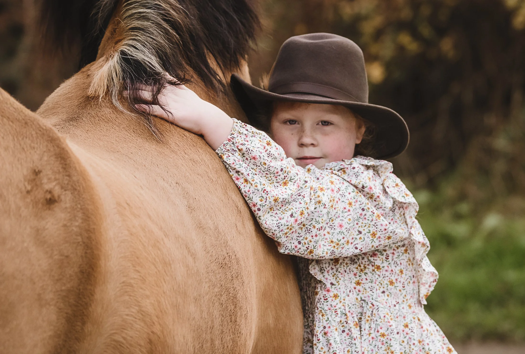 A young girl in a floral dress and a wide-brimmed hat hugging a brown horse outdoors.