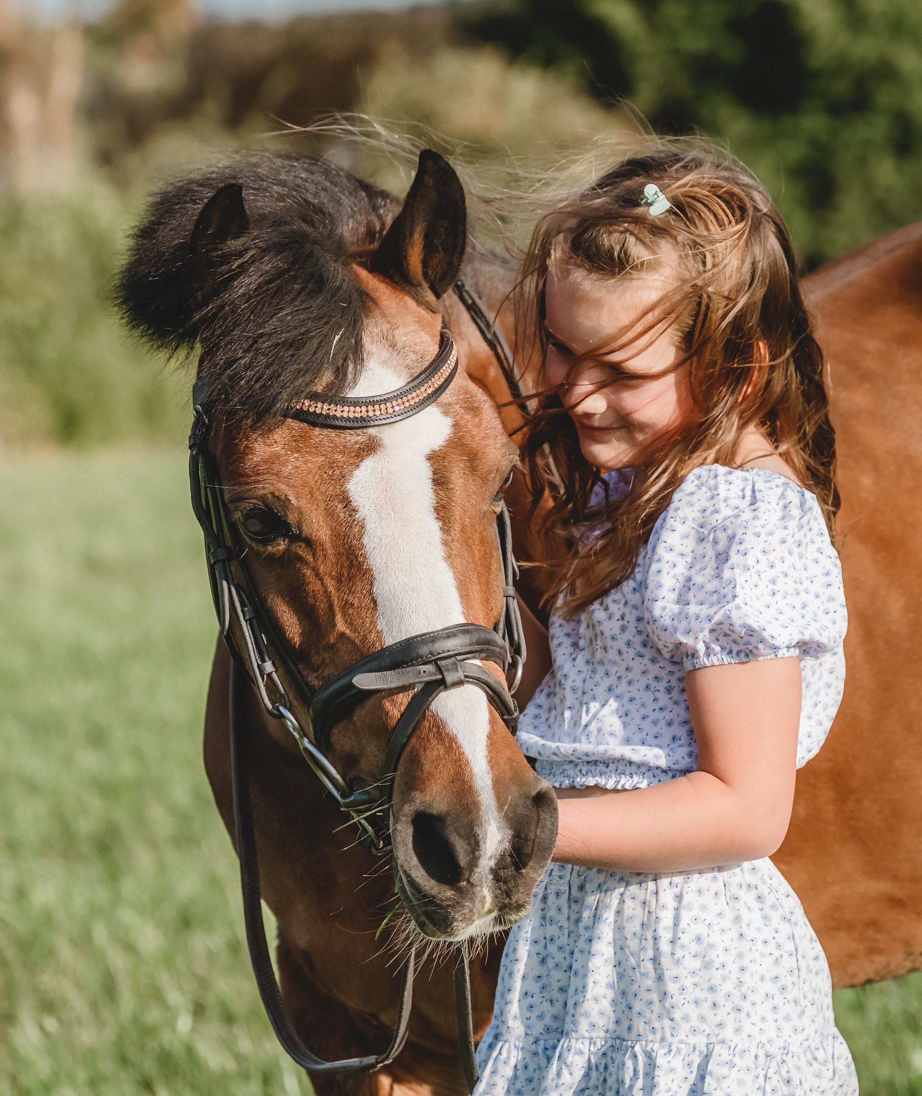 A young girl with brown hair and a blue hairclip, wearing a white dress with small blue flowers, is embracing a brown horse with a white stripe down its face. The girl is smiling and the horse has a bridle on, standing in a grassy outdoor field.