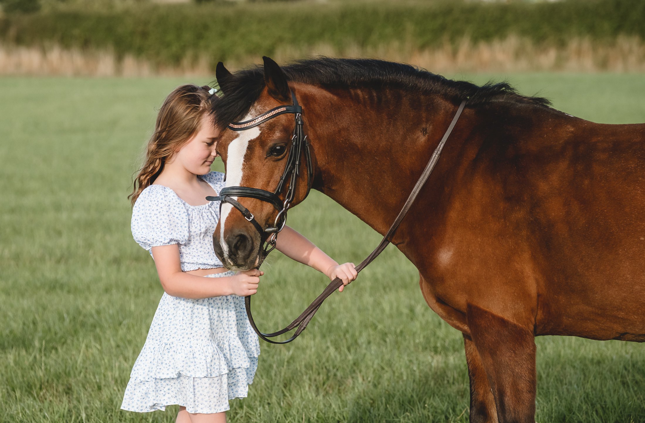 A young girl in a white dress with blue patterns stands on a grassy field, gently resting her forehead against a brown horse's forehead, holding the horse's reins with both hands.