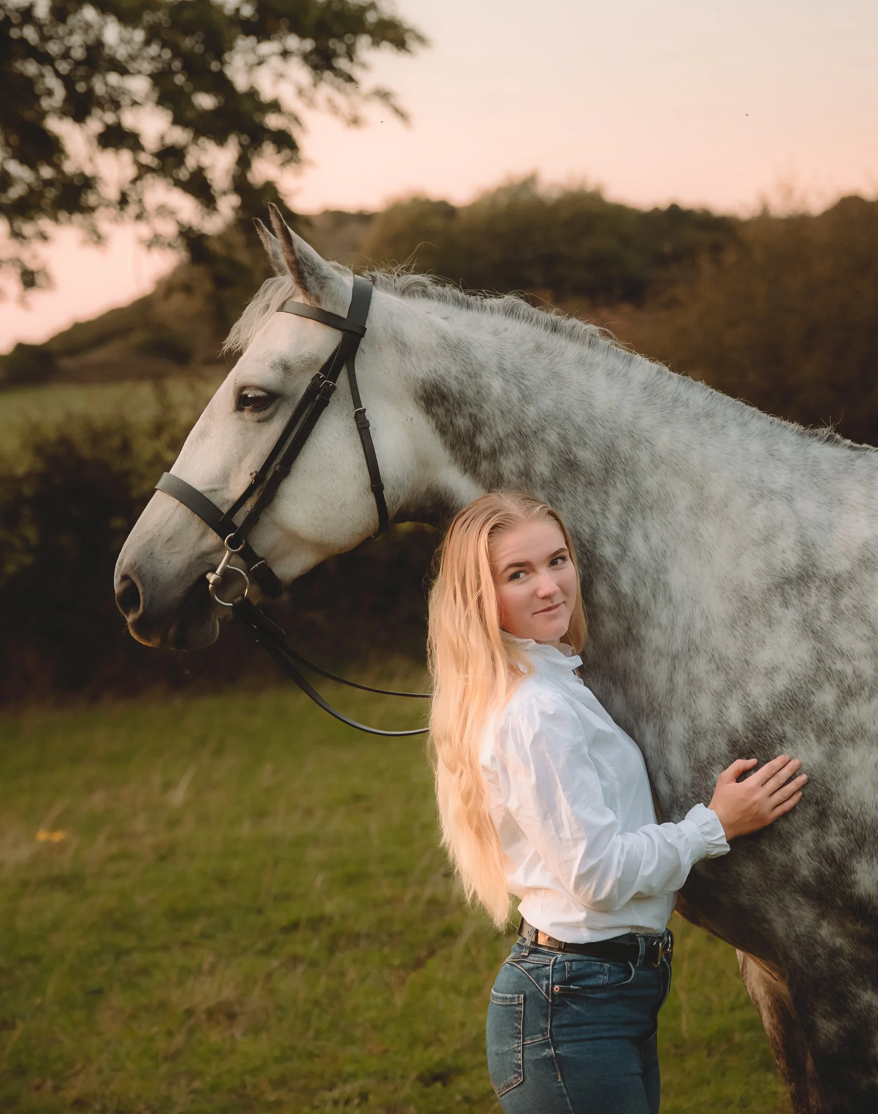 A young woman with long blonde hair stands beside a gray horse during sunset, holding the horse's neck and looking at the camera, with a background of trees and a pink sky.