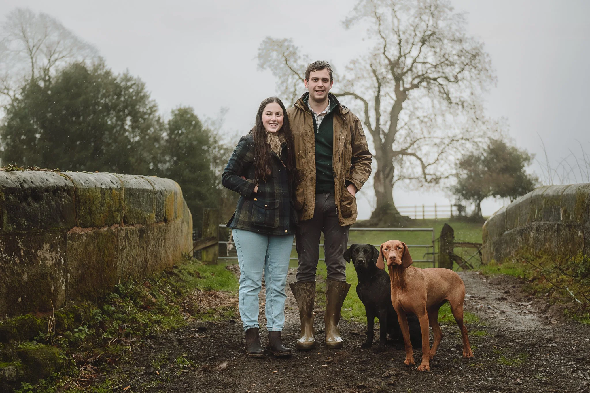 A young couple standing on a muddy path outdoors with two dogs, one black and one brown, on a misty day with leafless trees and moss-covered stone walls in the background.