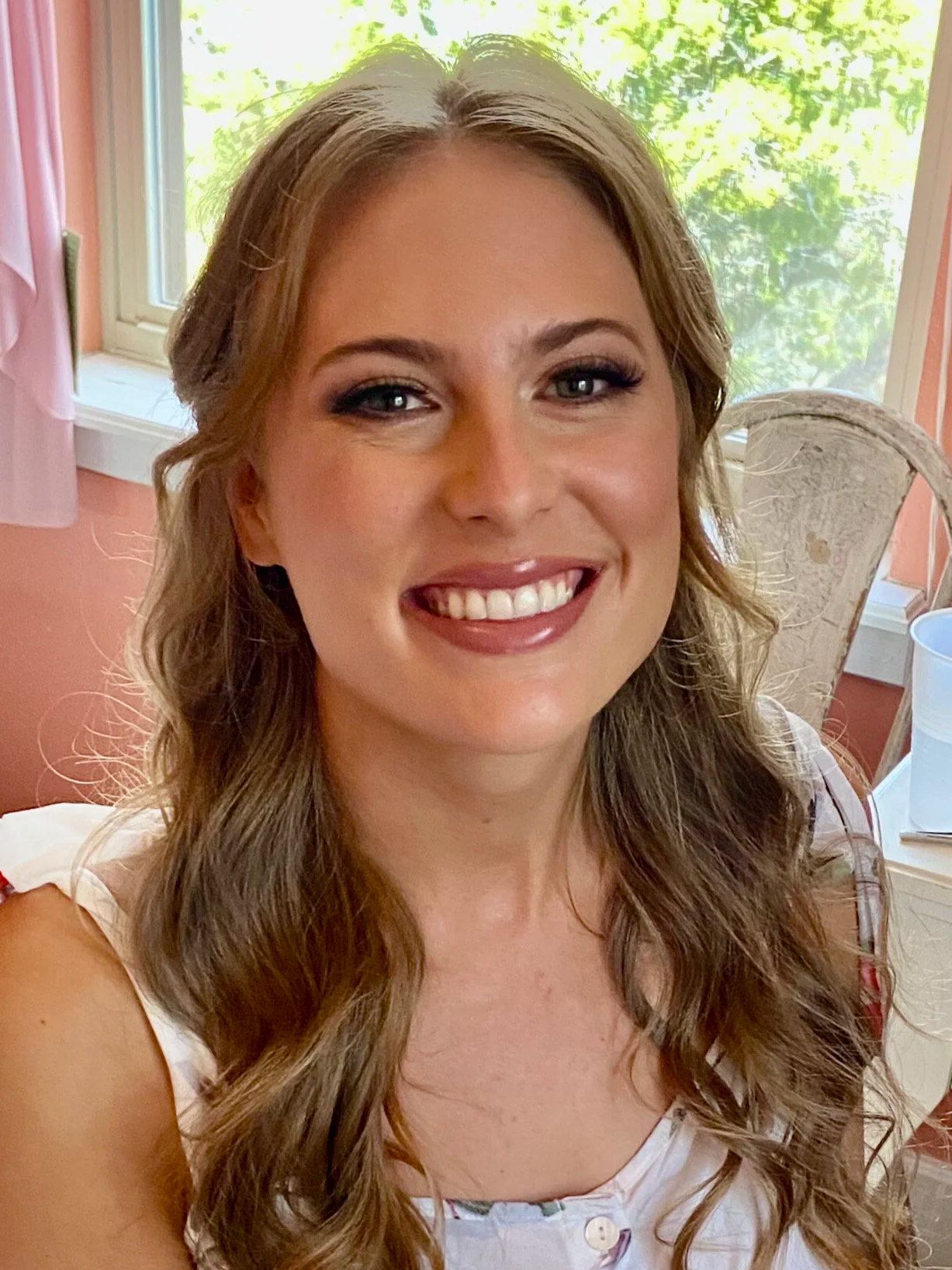 A smiling woman with wavy light brown hair sitting in a room with pink walls and floral curtains. Behind her, a window with greenery outside, a white table with various items, including a photo frame, cups, and bottles.