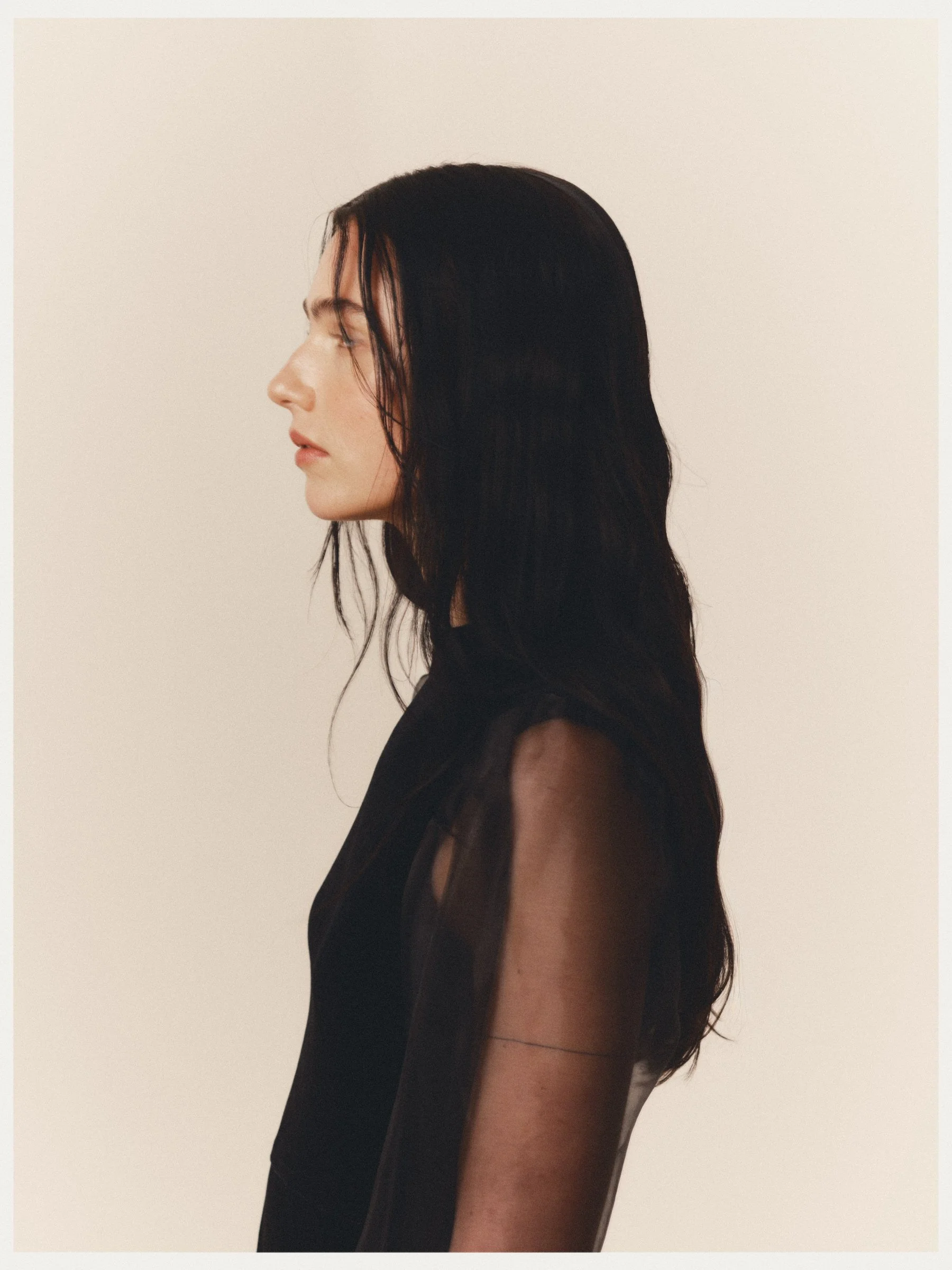 Side profile of a woman with long black hair, wearing a black sleeveless top, standing against a plain white background.
