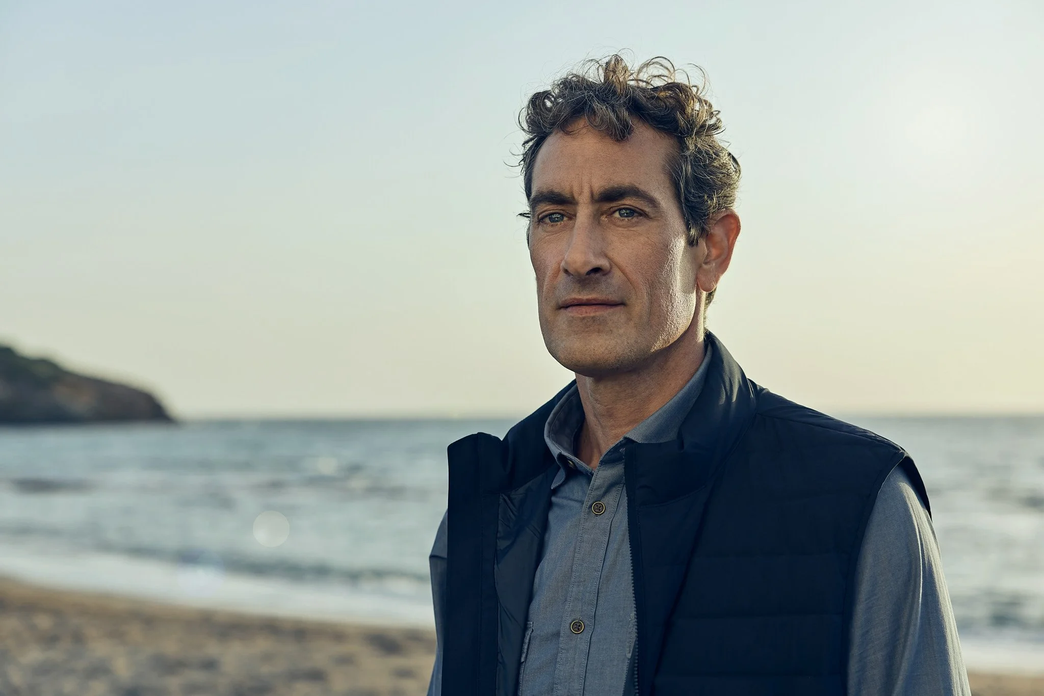 A middle-aged man with curly hair wearing a grey shirt and black vest, standing on a beach with the ocean and a small landmass in the background.