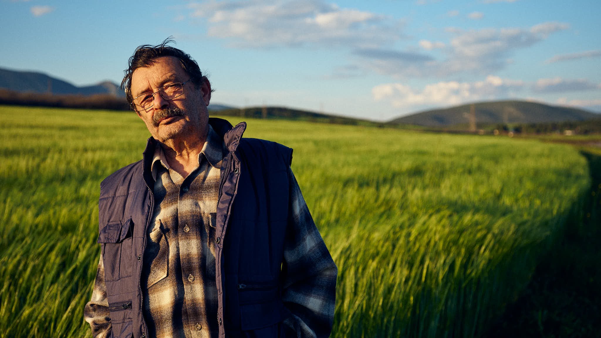 Older man with glasses, mustache, and beard standing in a green field during sunset, wearing a plaid shirt, vest, and jacket.