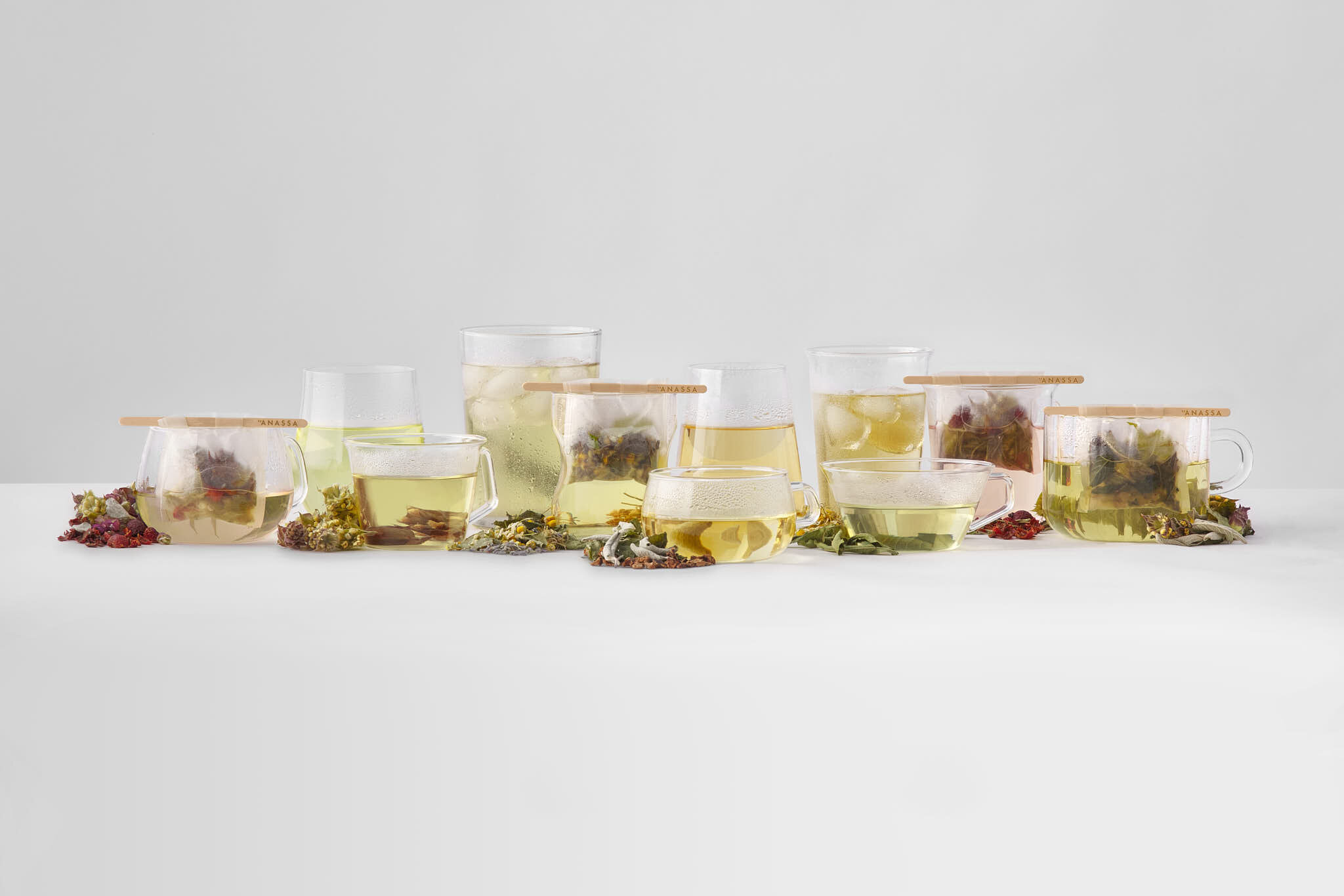 Several glass cups and mugs filled with herbal tea, surrounded by dried herbs and flower petals on a white surface with a plain white background.