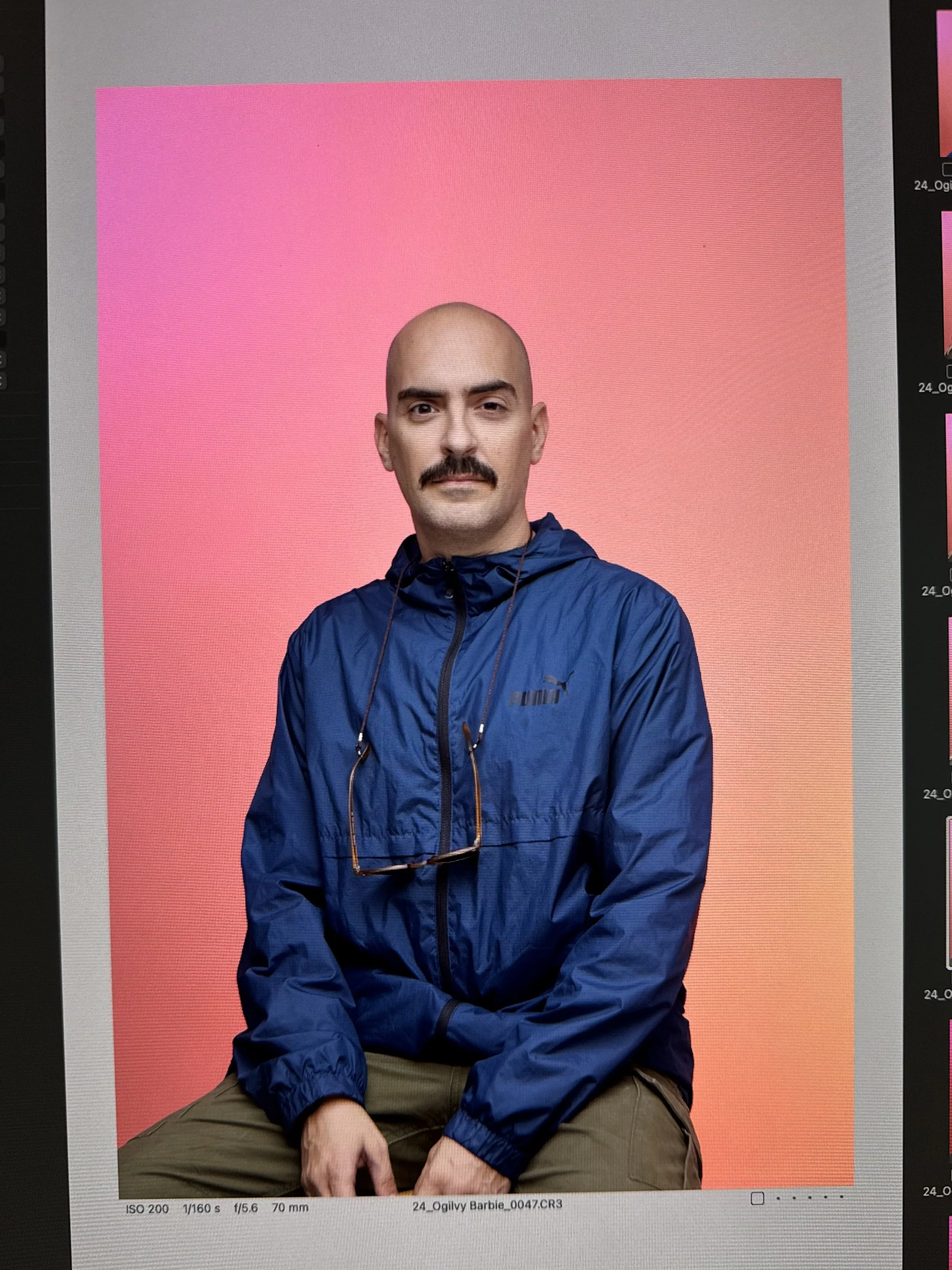 A man with a bald head, mustache, and beige skin wearing a blue jacket and khaki pants seated against a pink and orange gradient background.
