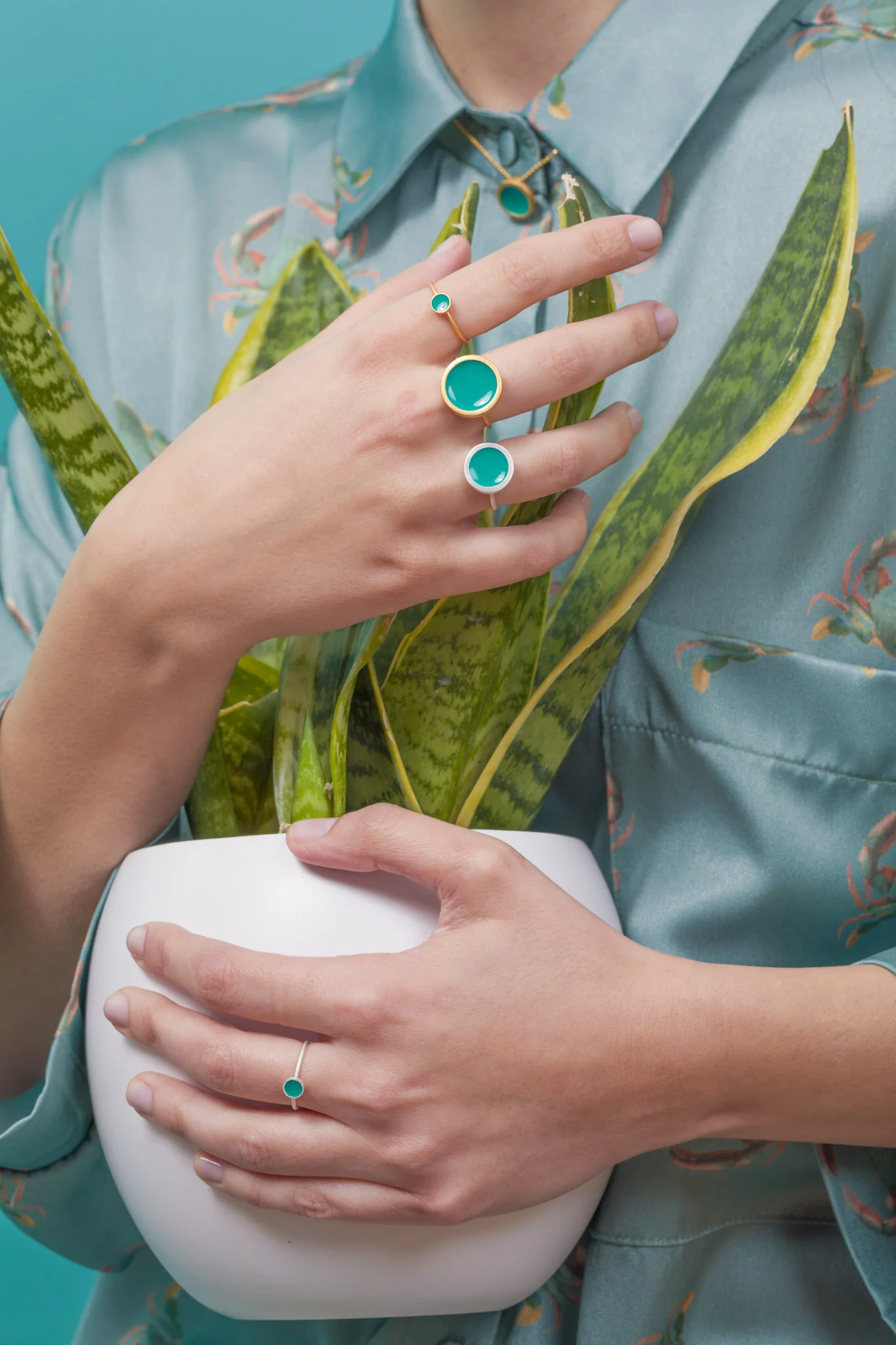 A person holds a white pot with green and yellow leaves, wearing jewelry with turquoise stones, including rings and necklaces, against a teal background.
