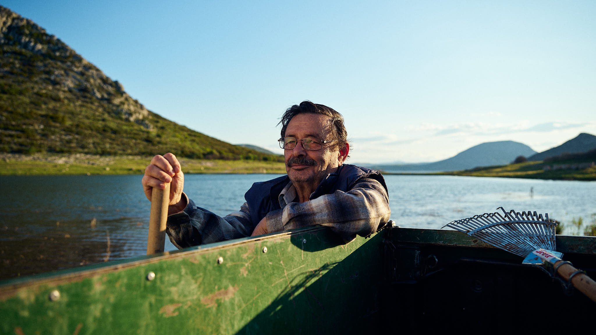 An older man with glasses and a beard smiling while sitting in a green canoe on a lake, holding a paddle. Mountains and a partly cloudy sky are in the background.