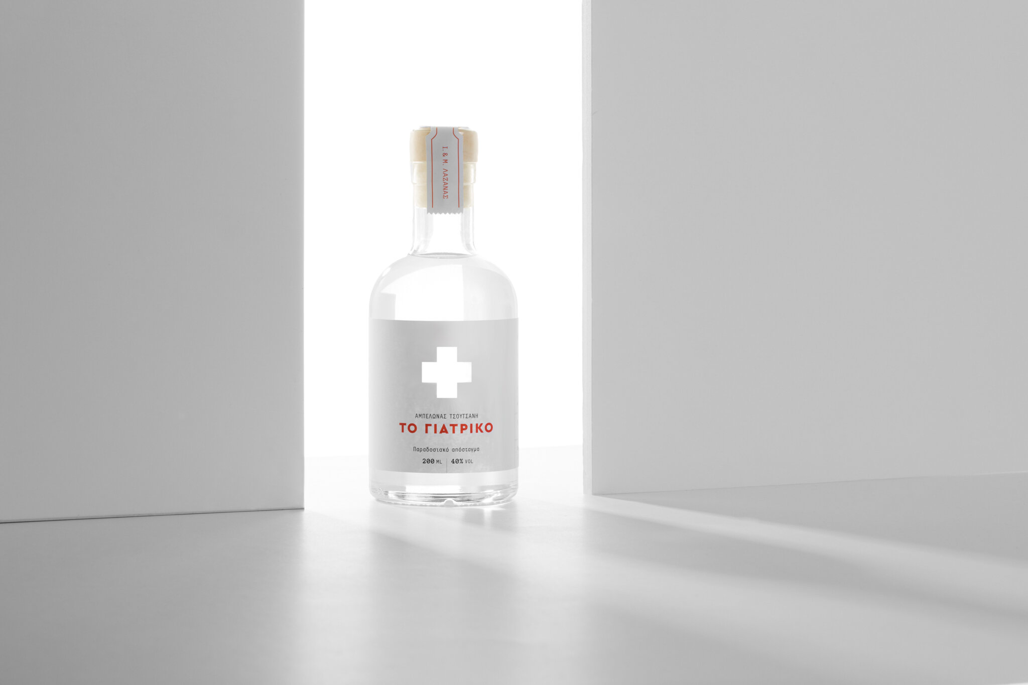 A clear glass bottle with Greek writing and a white cross symbol on the label, placed between two white walls with bright backlighting.