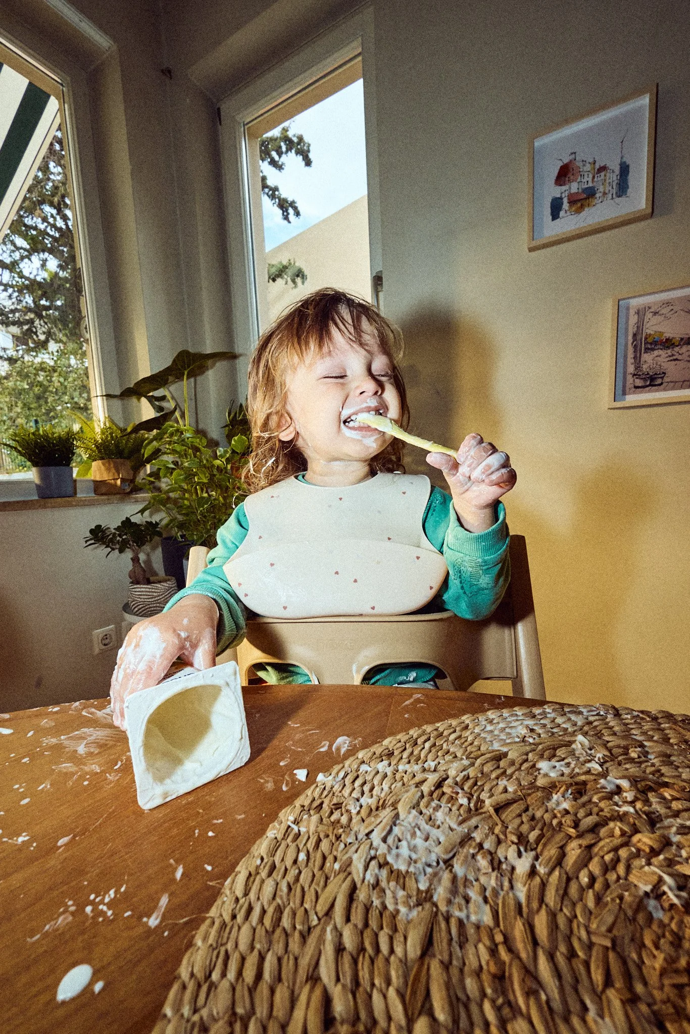 Young child with curly hair and a bib eating with a spoon at a wooden table, surrounded by spilled cream and a bread roll, in a room with plants and windows.