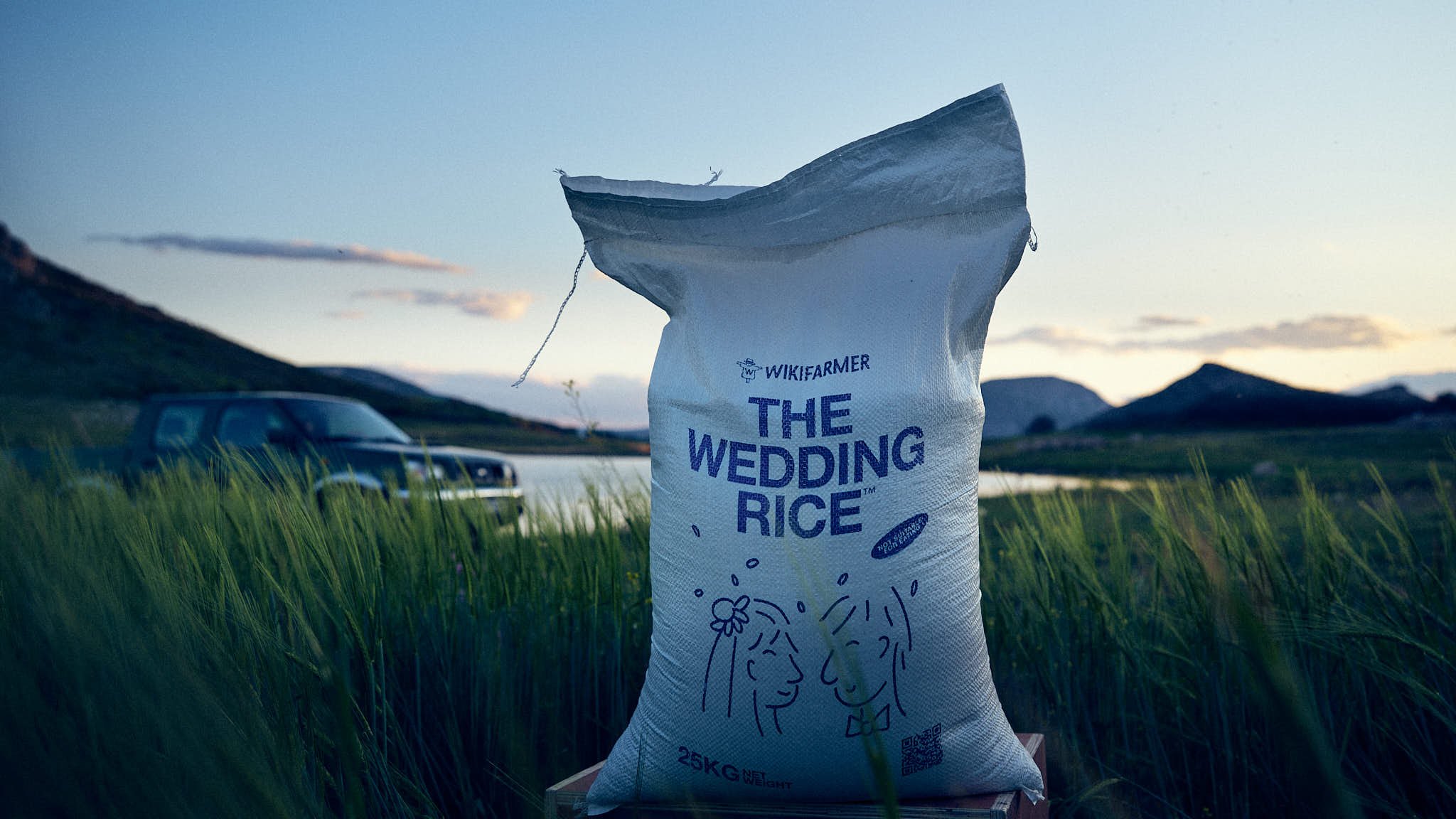 A bag of wedding rice labeled 'The Wedding Rice,' standing outdoors on a grassy area near a lake with mountains in the background during sunset.