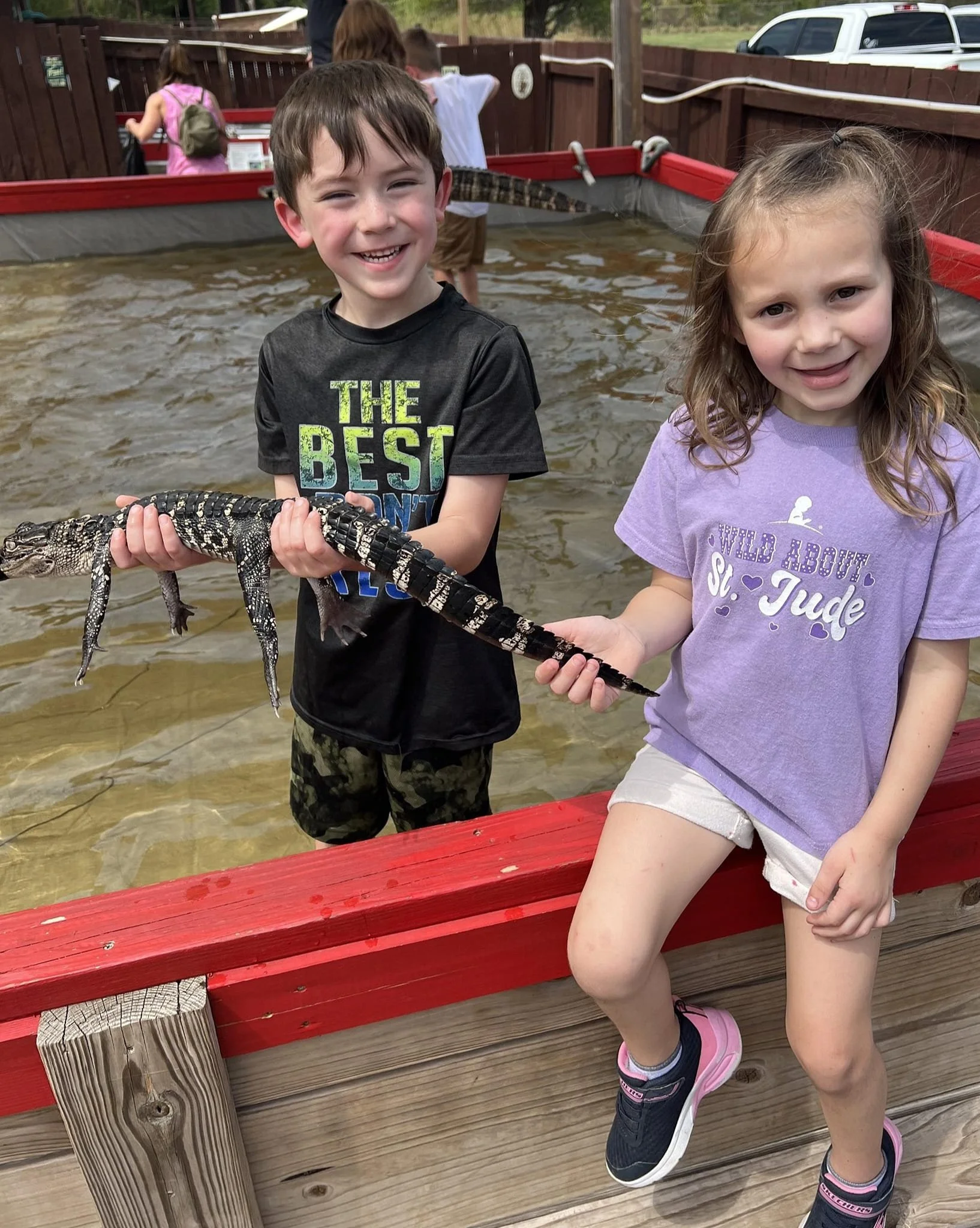 Two kids, a boy and a girl, holding a small crocodile or alligator in a red-fenced wildlife exhibit or pond, with other children in the background.