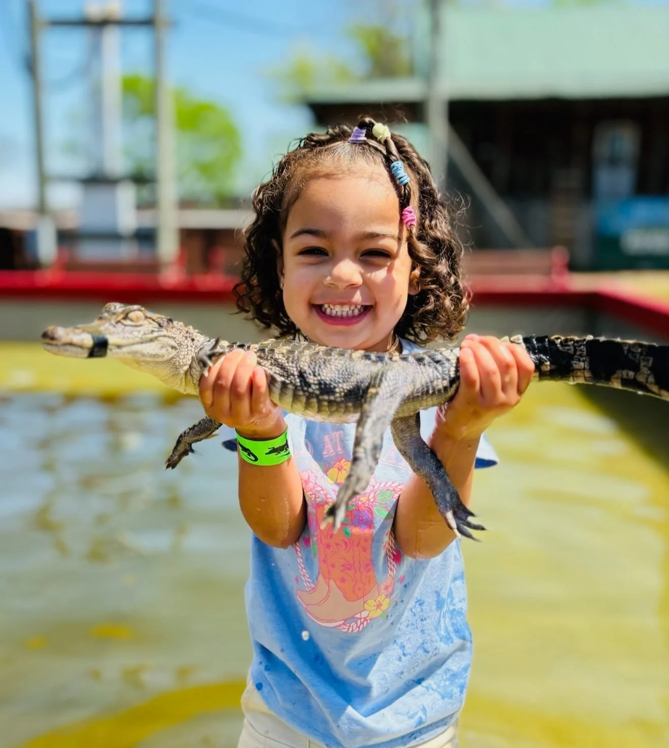 A young girl smiling and holding a small crocodile over water, outdoors on a sunny day.