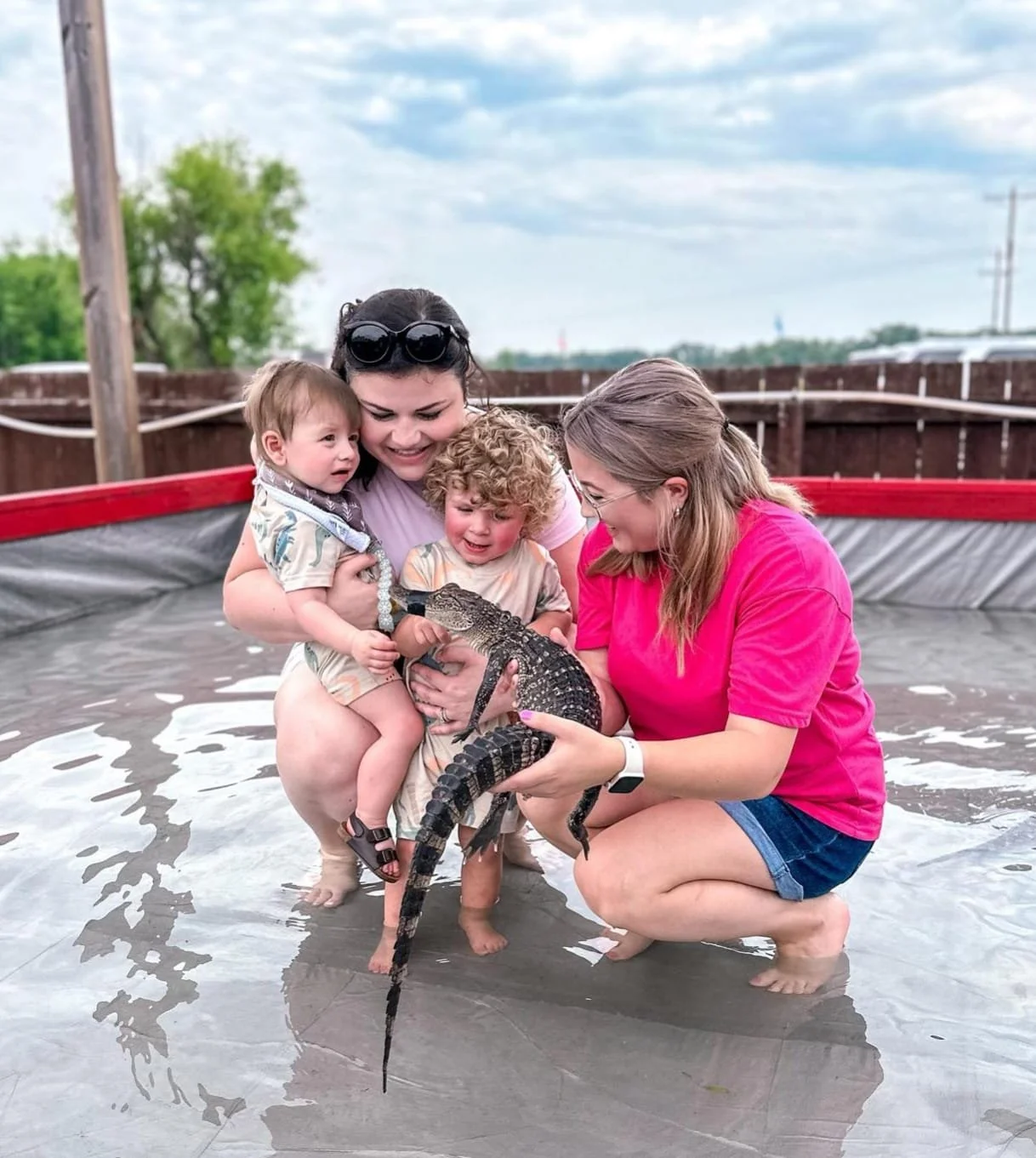 Four children and two women holding a young crocodile or alligator in a small pond or pool outdoors.