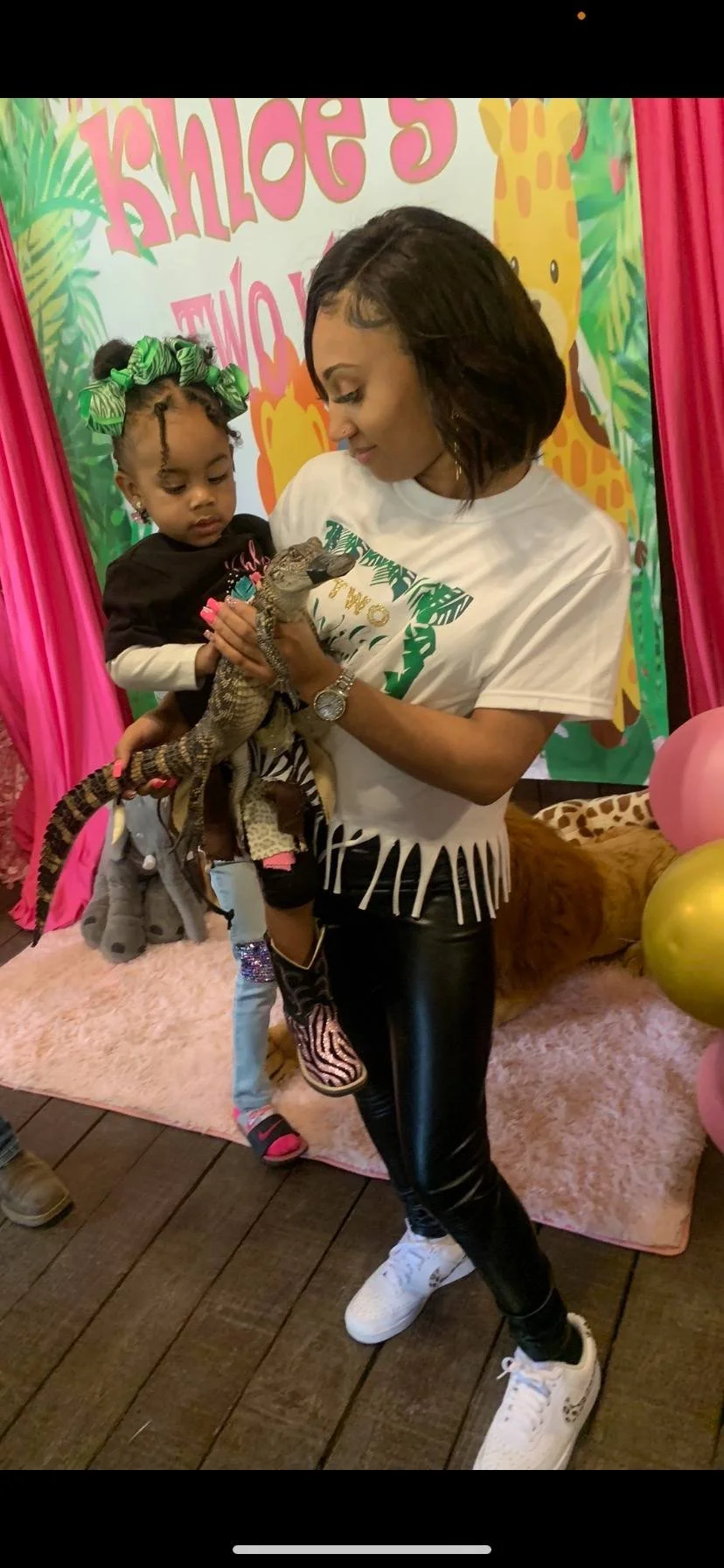 A woman and a young girl holding a small iguana at a jungle-themed birthday party with pink, yellow, and green decorations, including balloons and a backdrop with a giraffe and palm leaves.