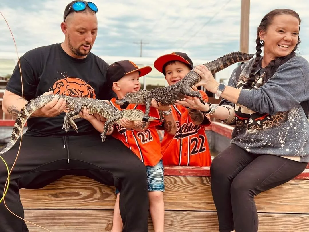 A family of four holding two small crocodiles outdoors, smiling and enjoying their time together.