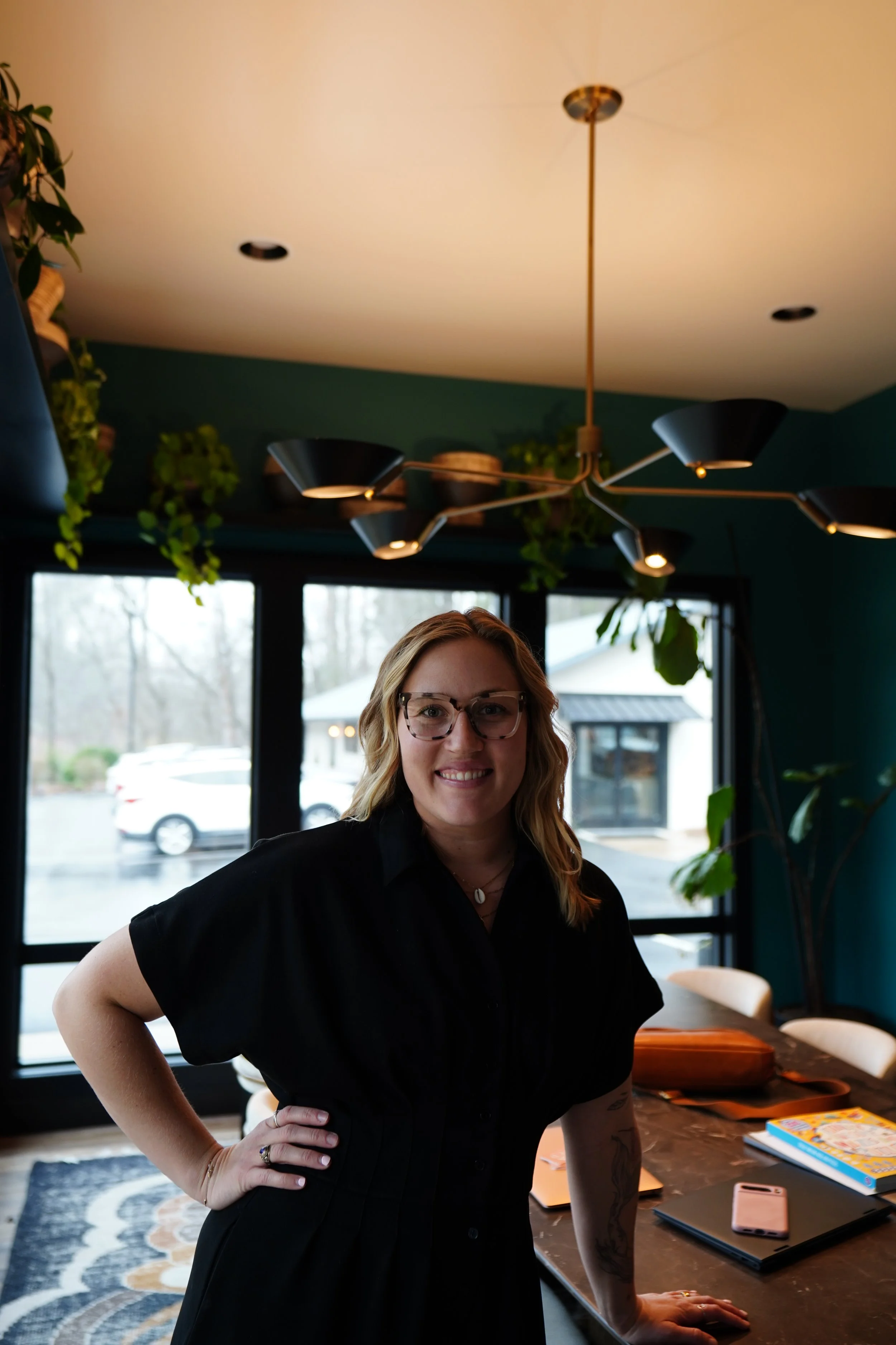 Woman with blonde hair and glasses smiling, standing with her hand on her hip in a modern dining area with large windows, hanging light fixture, and plants.