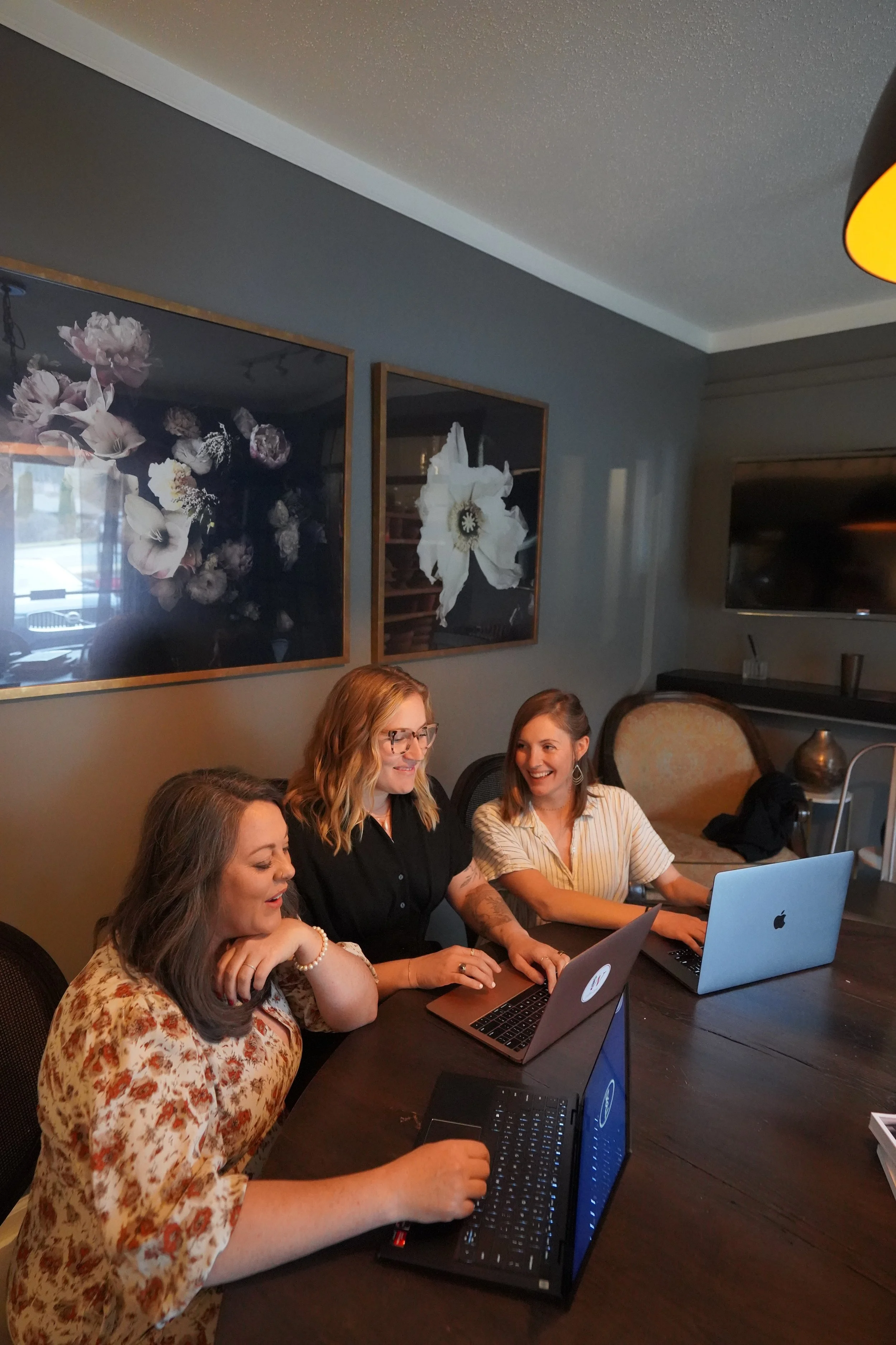 Three women sitting at a dark wooden table with laptops, smiling and chatting in a cozy room with floral artwork on the wall