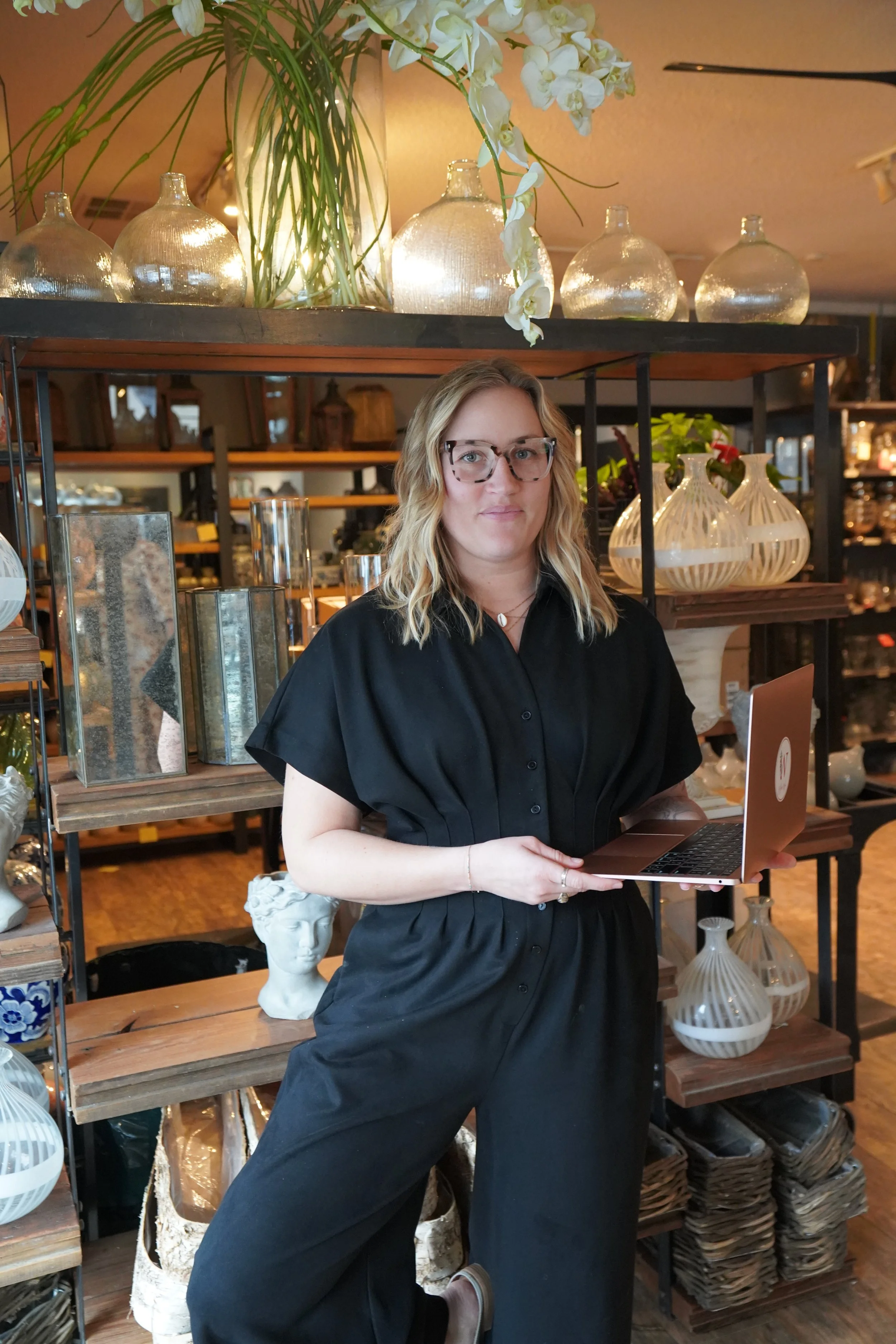 A woman with blond wavy hair, glasses, and a black outfit standing in a home decor store, holding a laptop, surrounded by decorative vases and statues.
