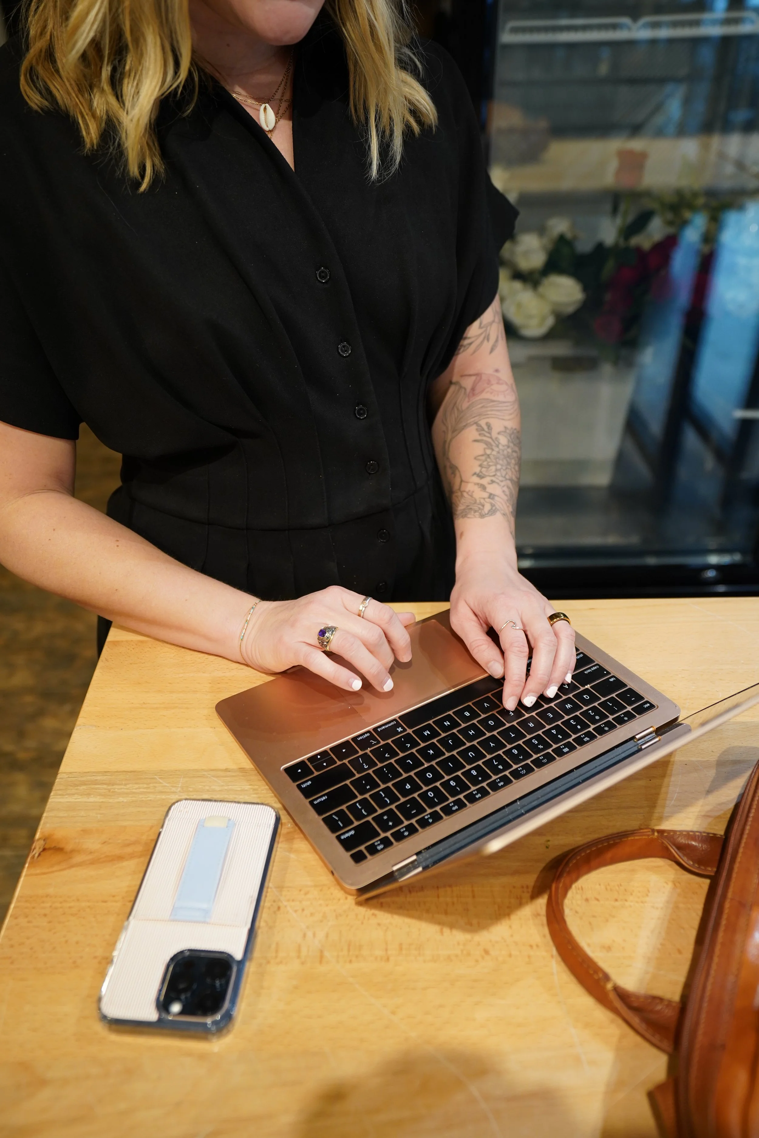 A woman with blonde hair, wearing a black dress with short sleeves, using a rose gold MacBook laptop on a wooden table. She has tattoos on her right arm, multiple rings on her fingers, and a shell necklace. A smartphone with a clear case and a white card inside is on the table.