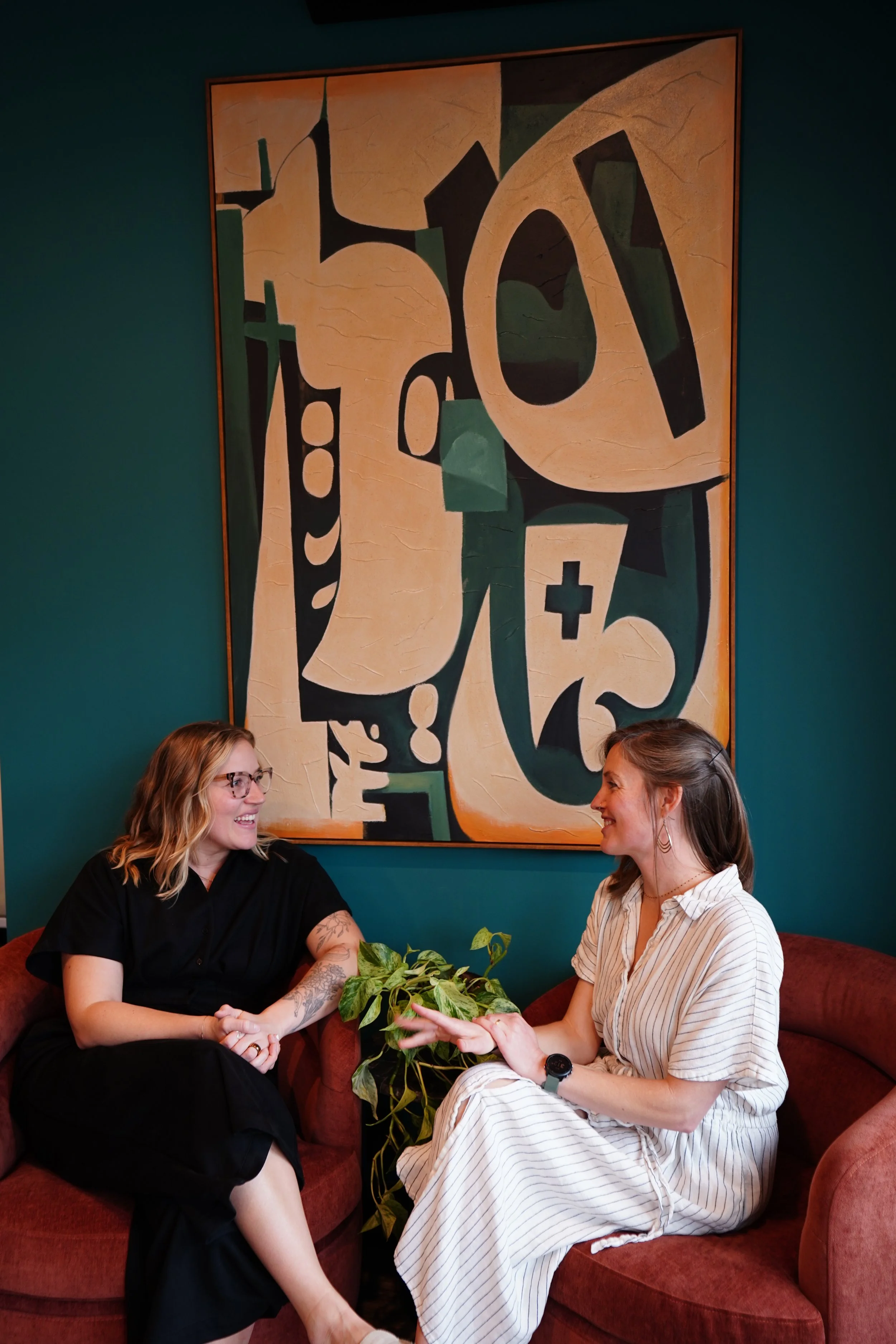 Two women sitting on a red sofa in a room, having a conversation and smiling at each other, with a large abstract painting on the teal wall behind them and a green plant between them.