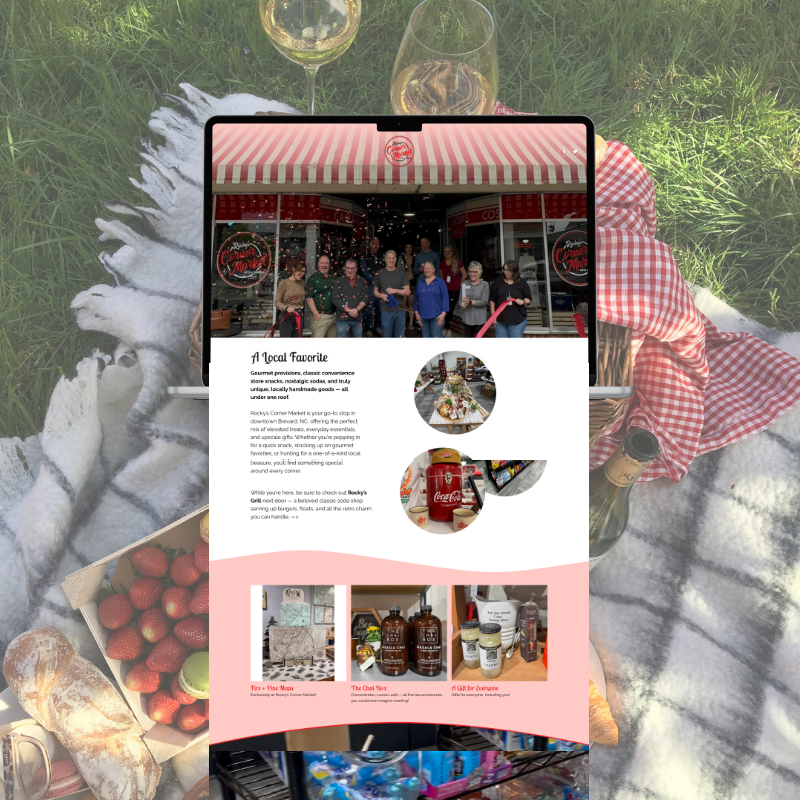 A tablet displaying a webpage about Rocky's Corner Market, a local grocery store, placed on a picnic blanket outdoors. The picnic blanket has a white furry texture with a red and white checked cloth and a glass of white wine, a bottle, and a box of strawberries nearby. The background is green grass.