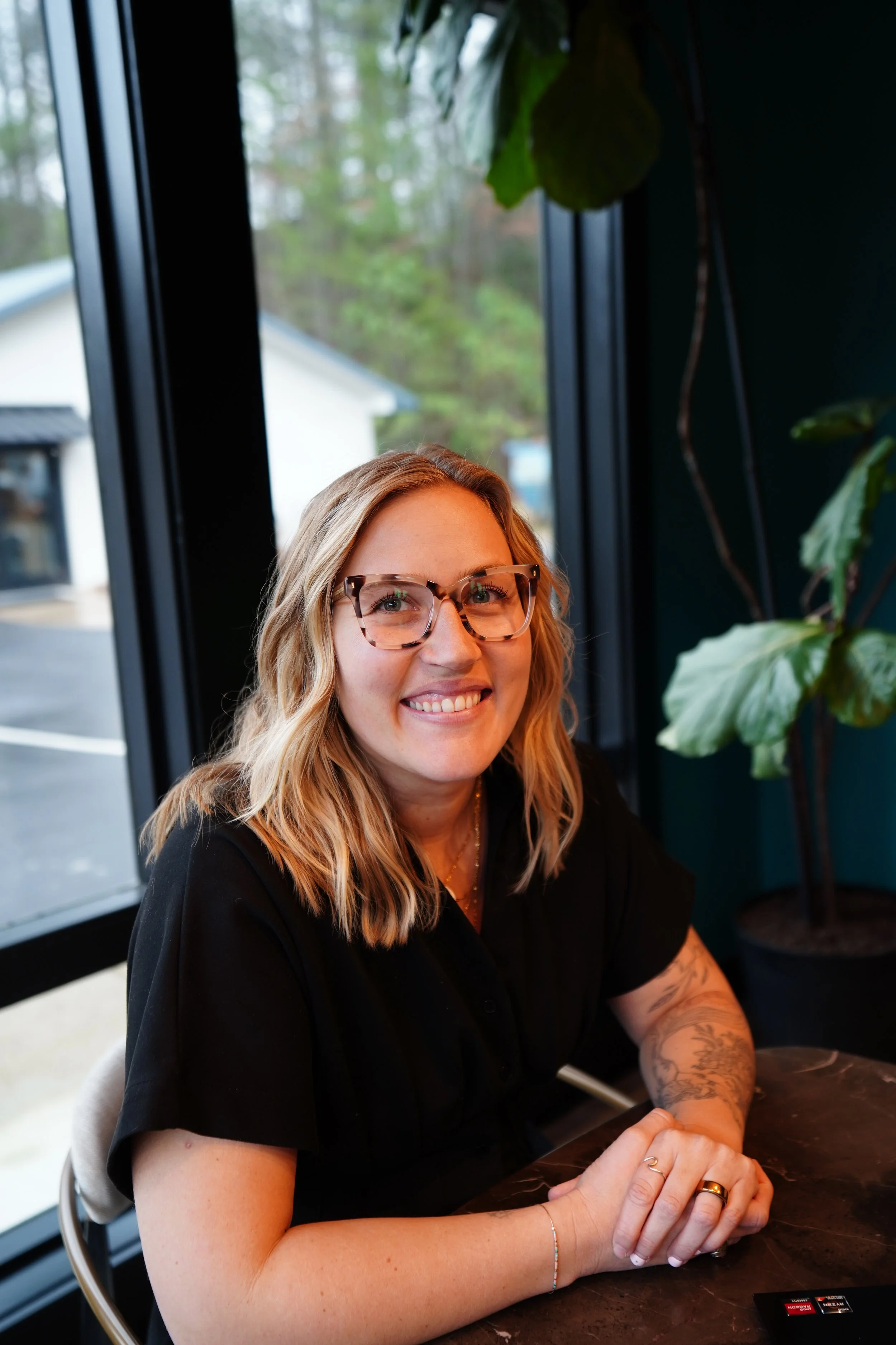 A woman with glasses and blonde hair smiling, sitting at a table in a cafe near a large window with plants outside.