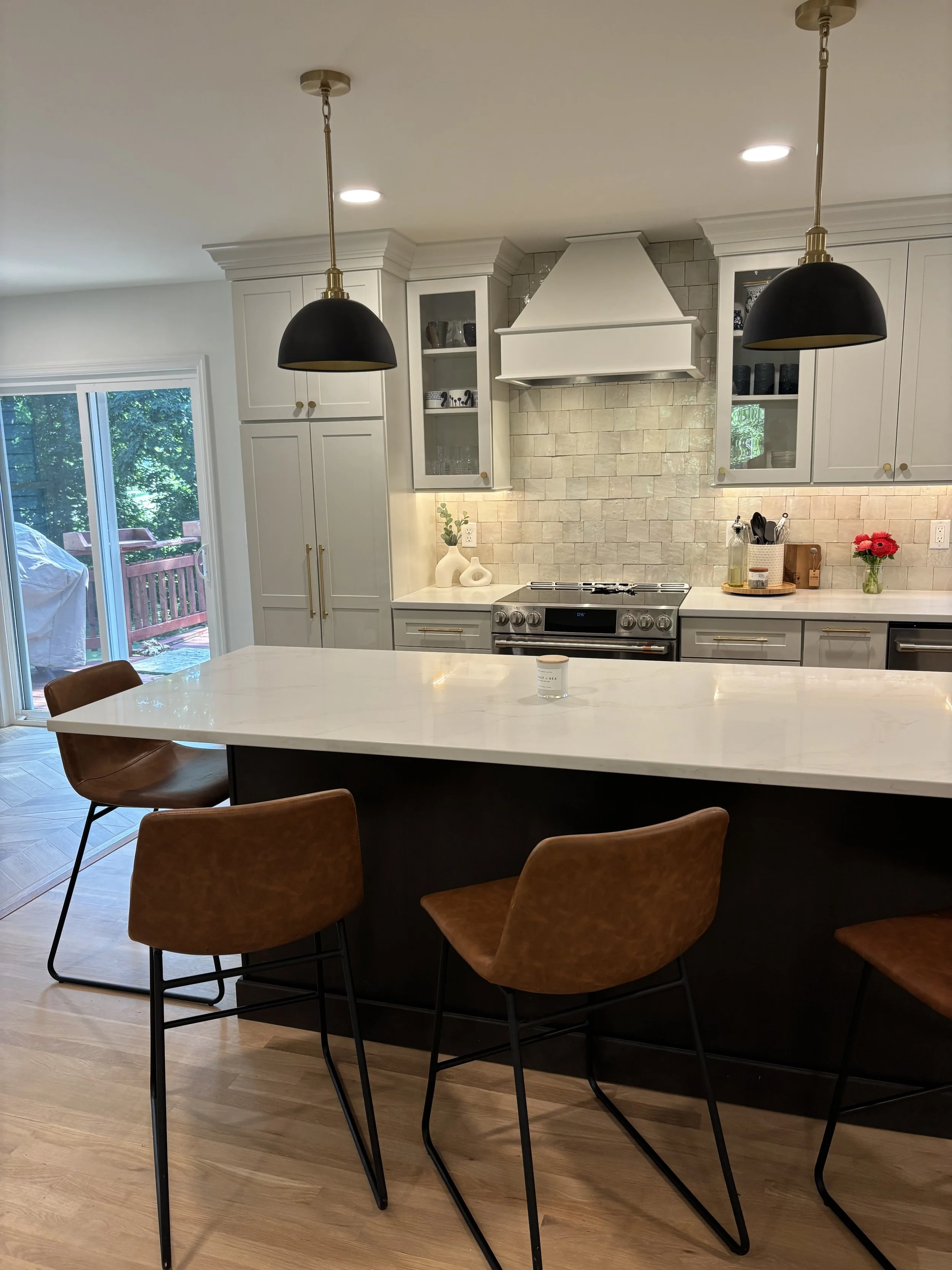 Modern kitchen with white cabinetry, a large island with a white countertop, and three brown bar stools with black legs. There are black pendant lights hanging above the island, and a sliding glass door leading to a deck outside. The area is well-lit