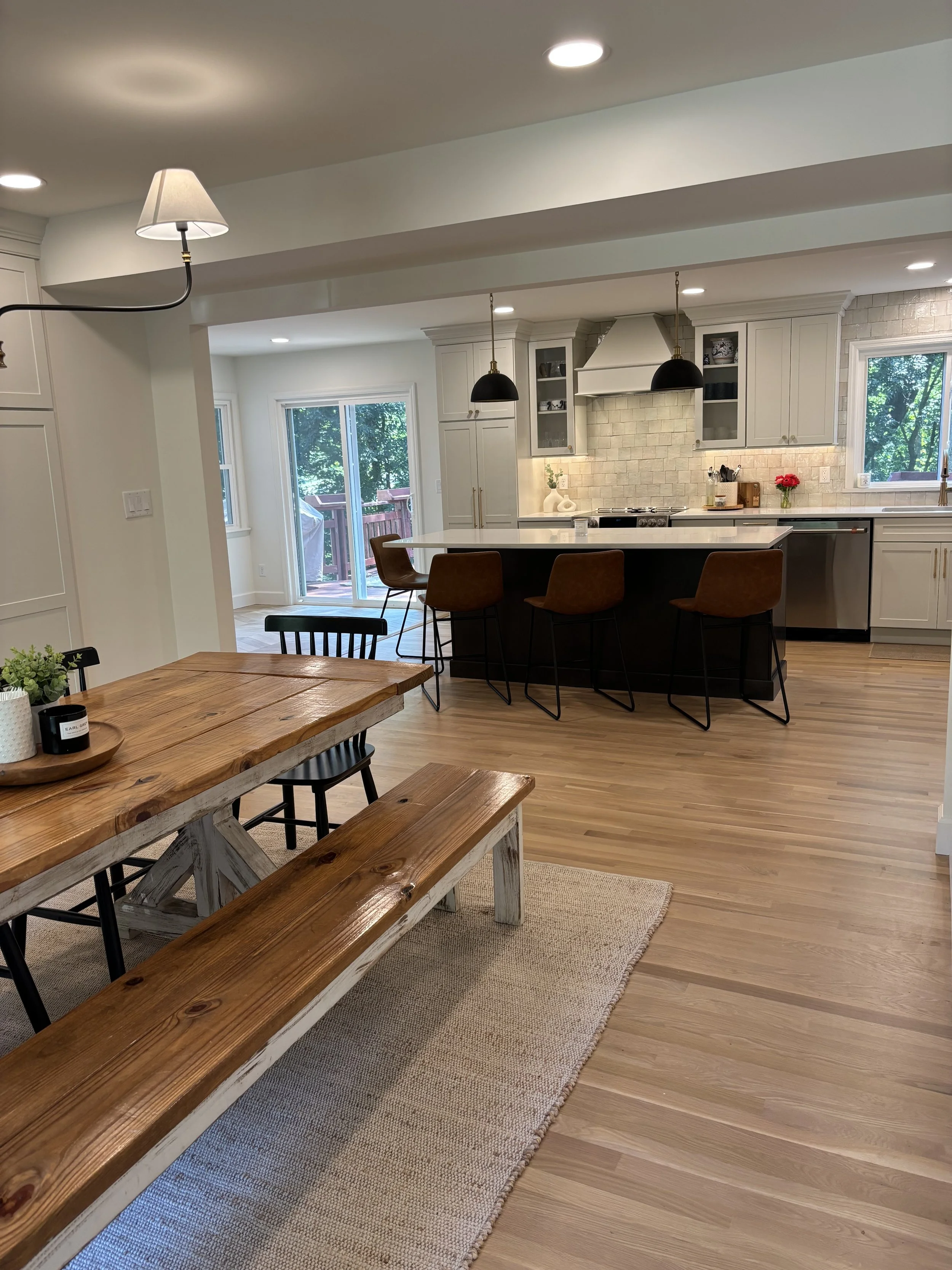 Open-concept kitchen and dining area with wooden dining table, benches, and chairs, white cabinets, island with barstools, pendant lights, and large windows with natural light.