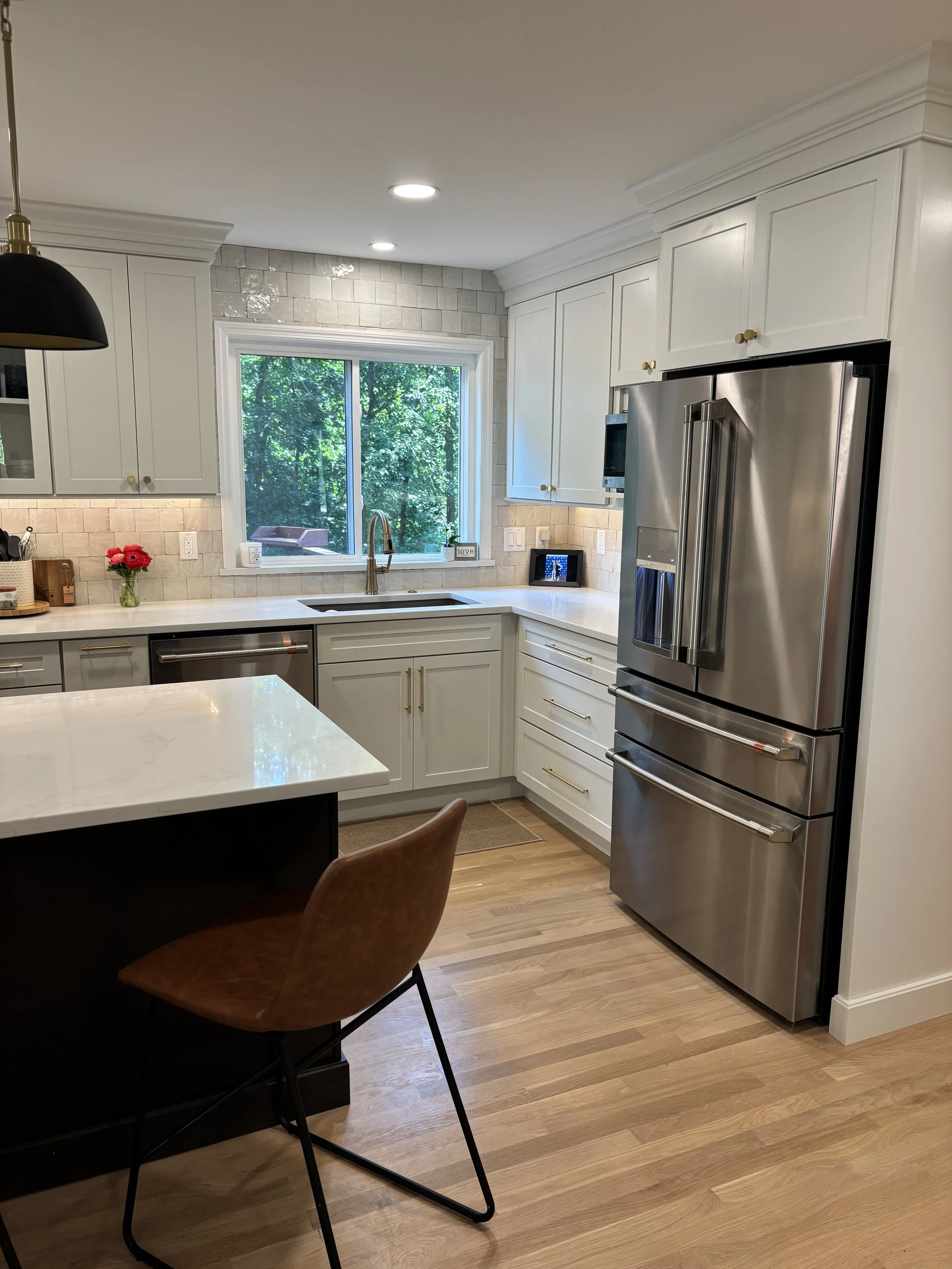 A modern kitchen with white cabinets, a large stainless steel refrigerator, a window overlooking greenery, and a marble-topped island with a brown chair.