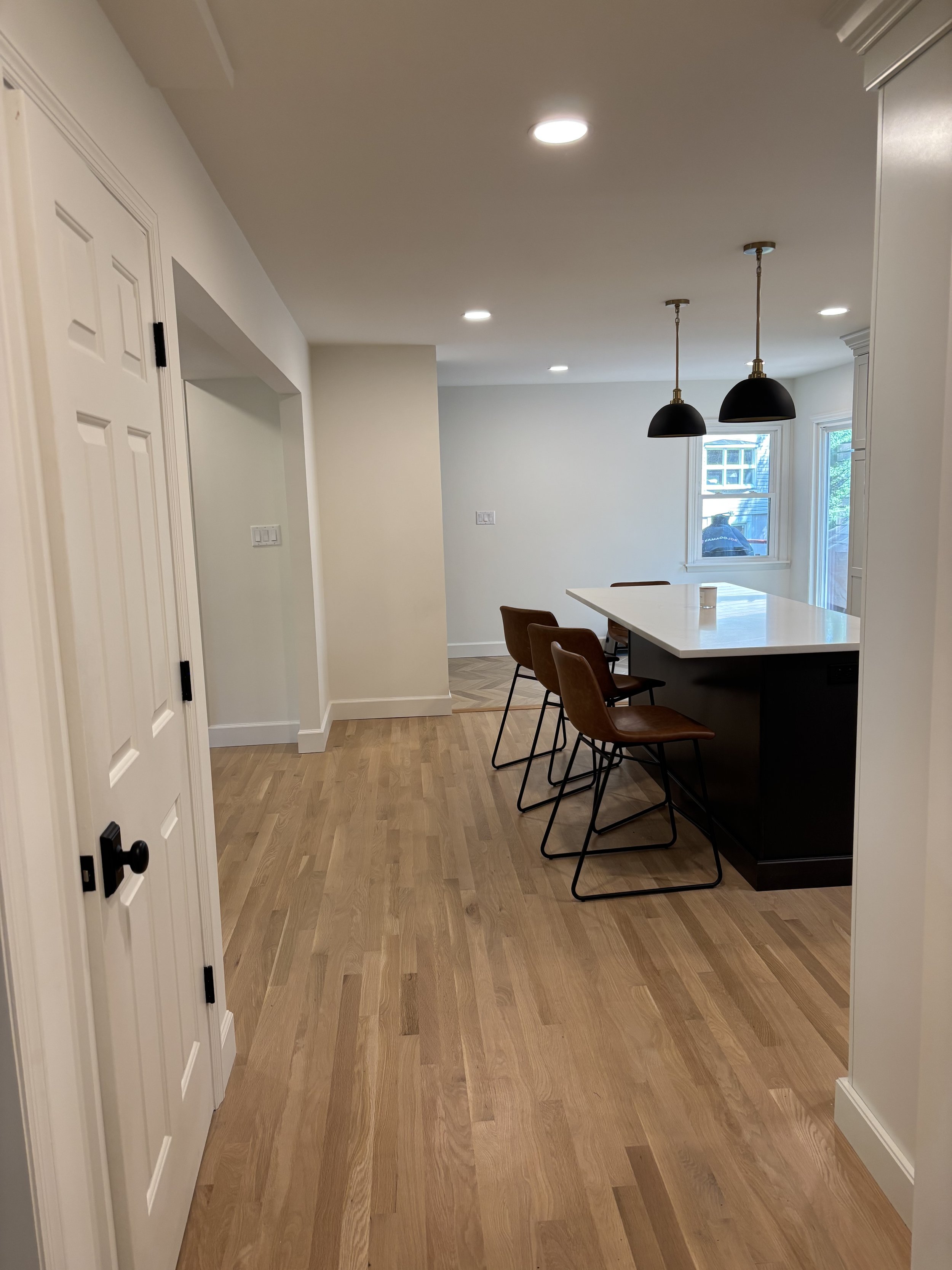 Modern kitchen with a large island, three brown chairs, pendant lights, hardwood floors, and a window showing a house outside.