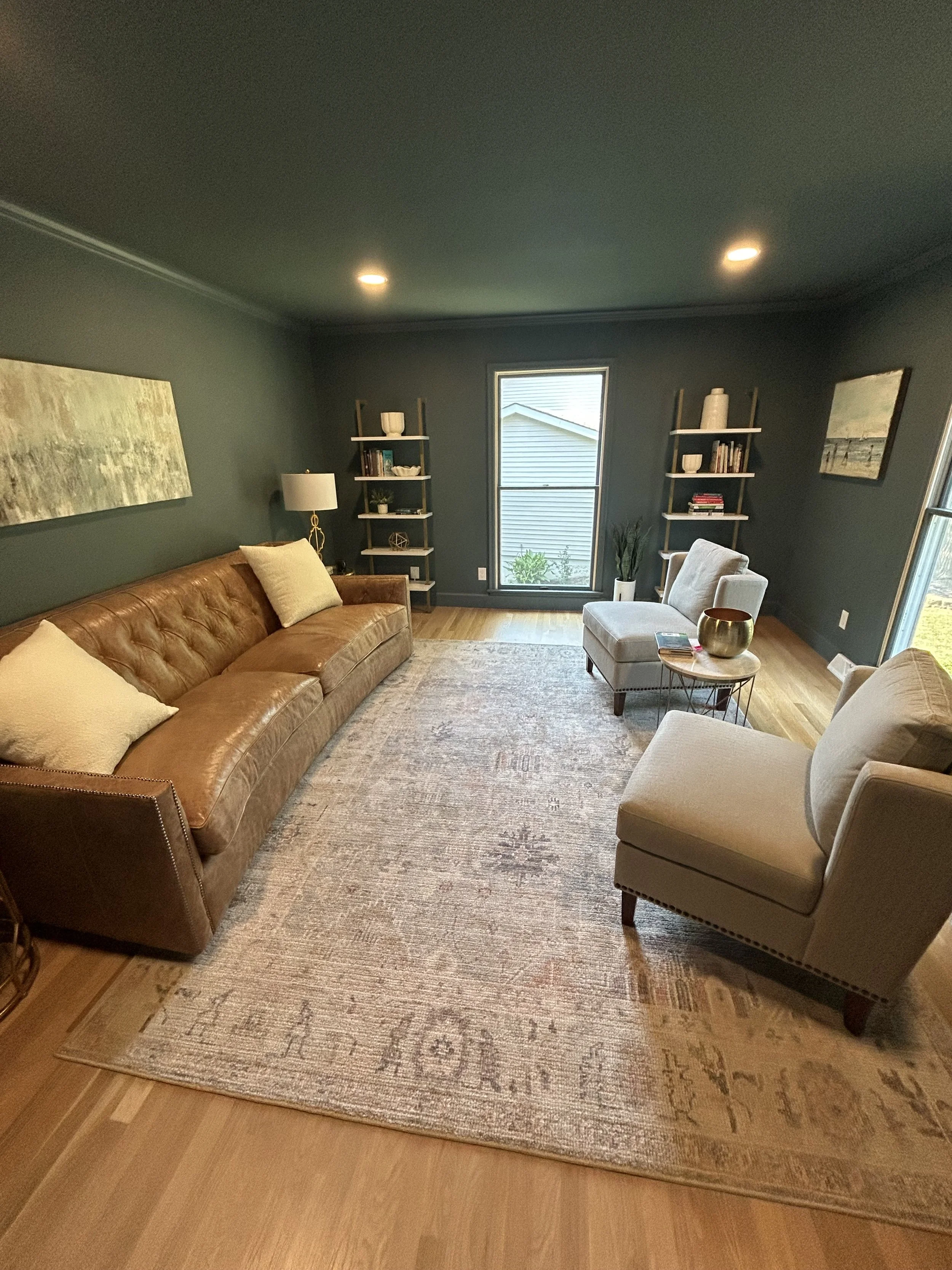 Living room with dark teal walls, hardwood flooring, a large brown leather sofa with pillows, a patterned area rug, a light gray armchair and ottoman, two wall-mounted shelves with books and decor, a window with a view of the outside, and a painting 