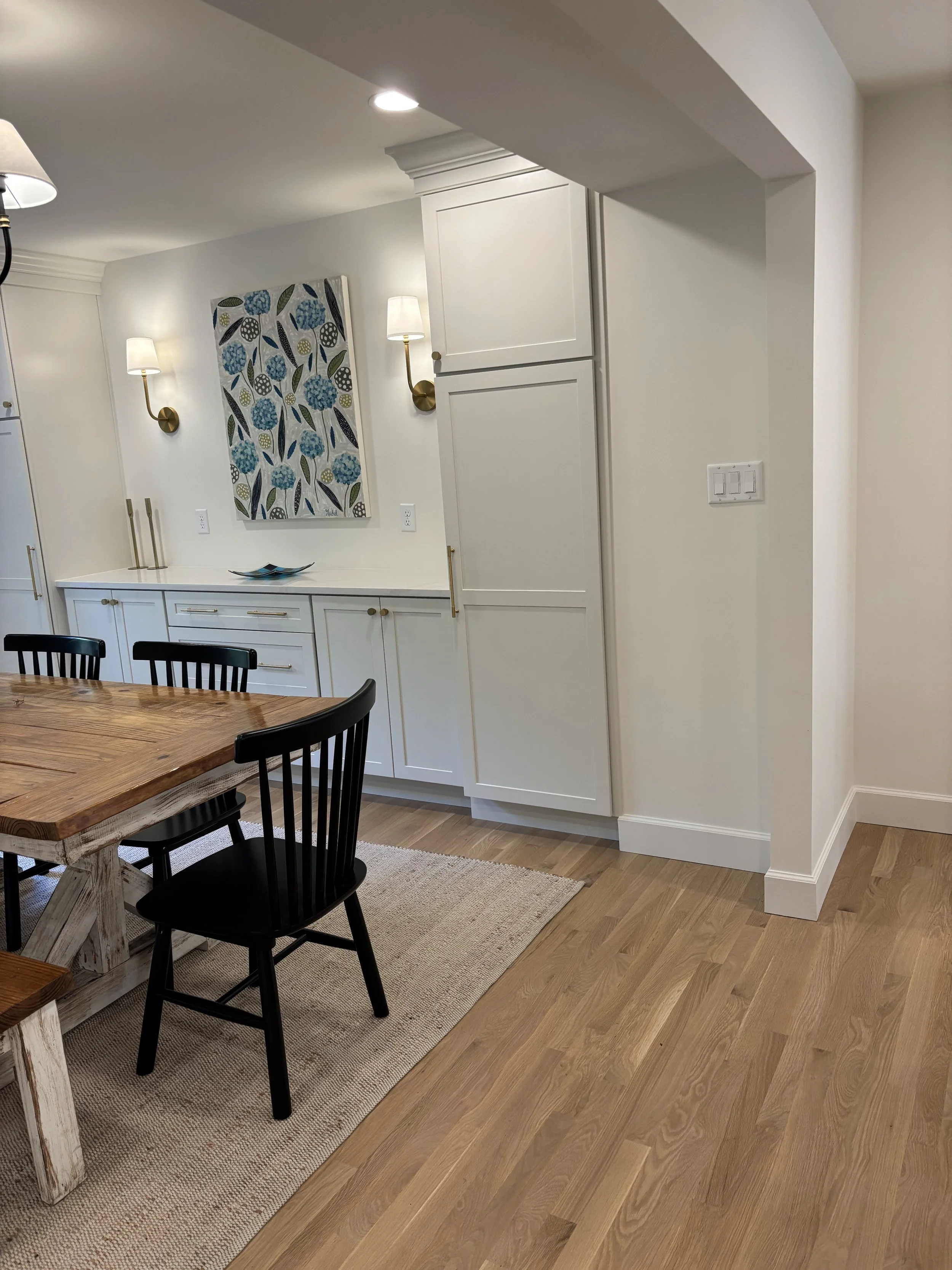 Dining area with a wooden table, black chairs, and white cabinetry, with wall-mounted lights, artwork, and hardwood flooring.